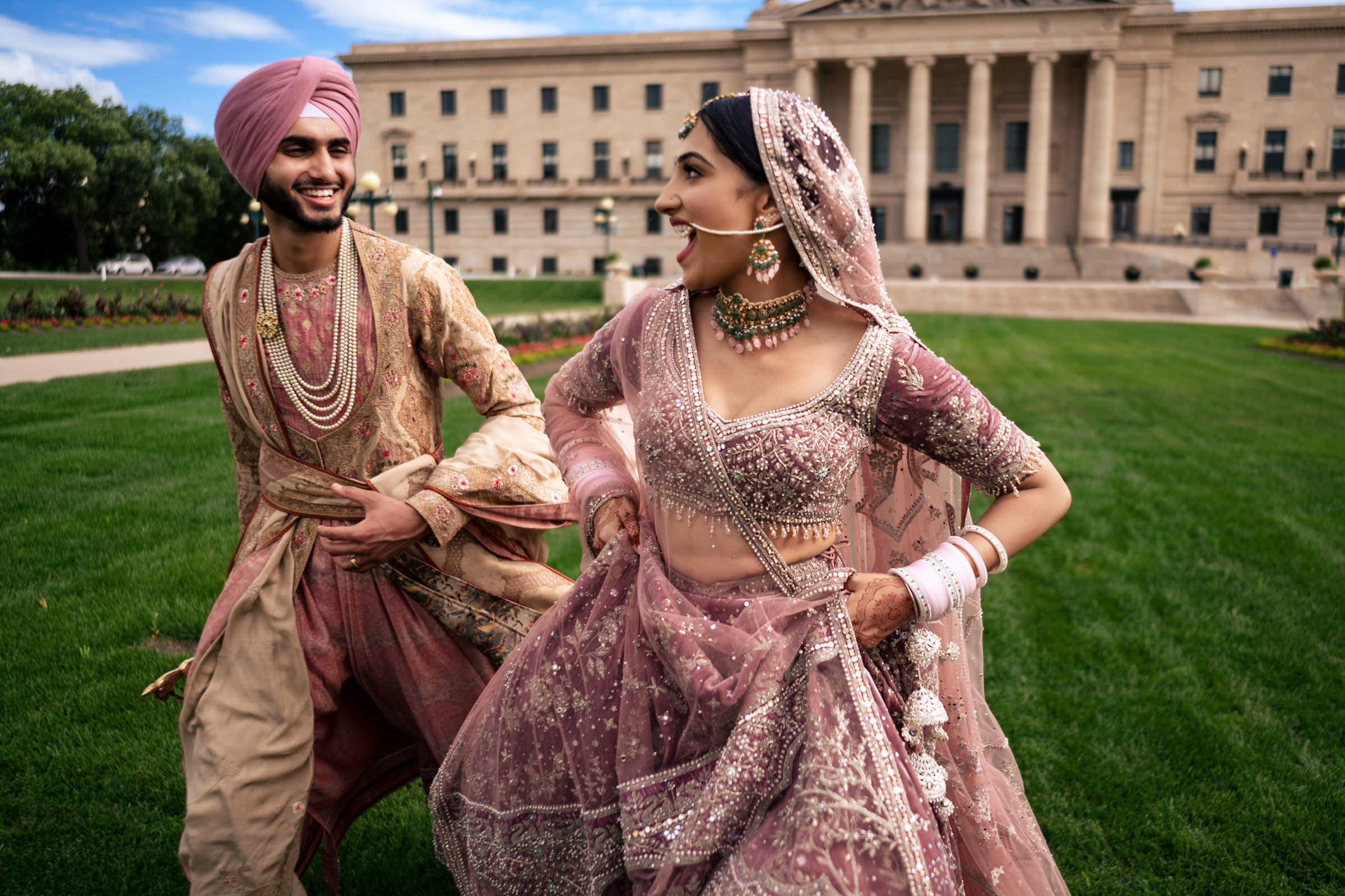 A joyful couple in traditional Indian attire, with a man in a pink turban and an embellished outfit, and a woman in a detailed pink lehenga. Captured among their favorite wedding photos, they laugh and run on a lush green lawn before a grand building.