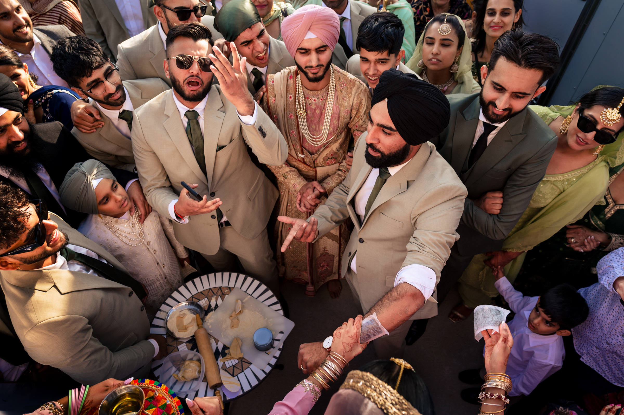 A group in colorful traditional attire gathers around a table during a lively celebration. Men in beige suits and a groom with a pink turban take center stage, with women and children surrounding them. Various items adorn the table, capturing the essence of their favorite wedding photos.