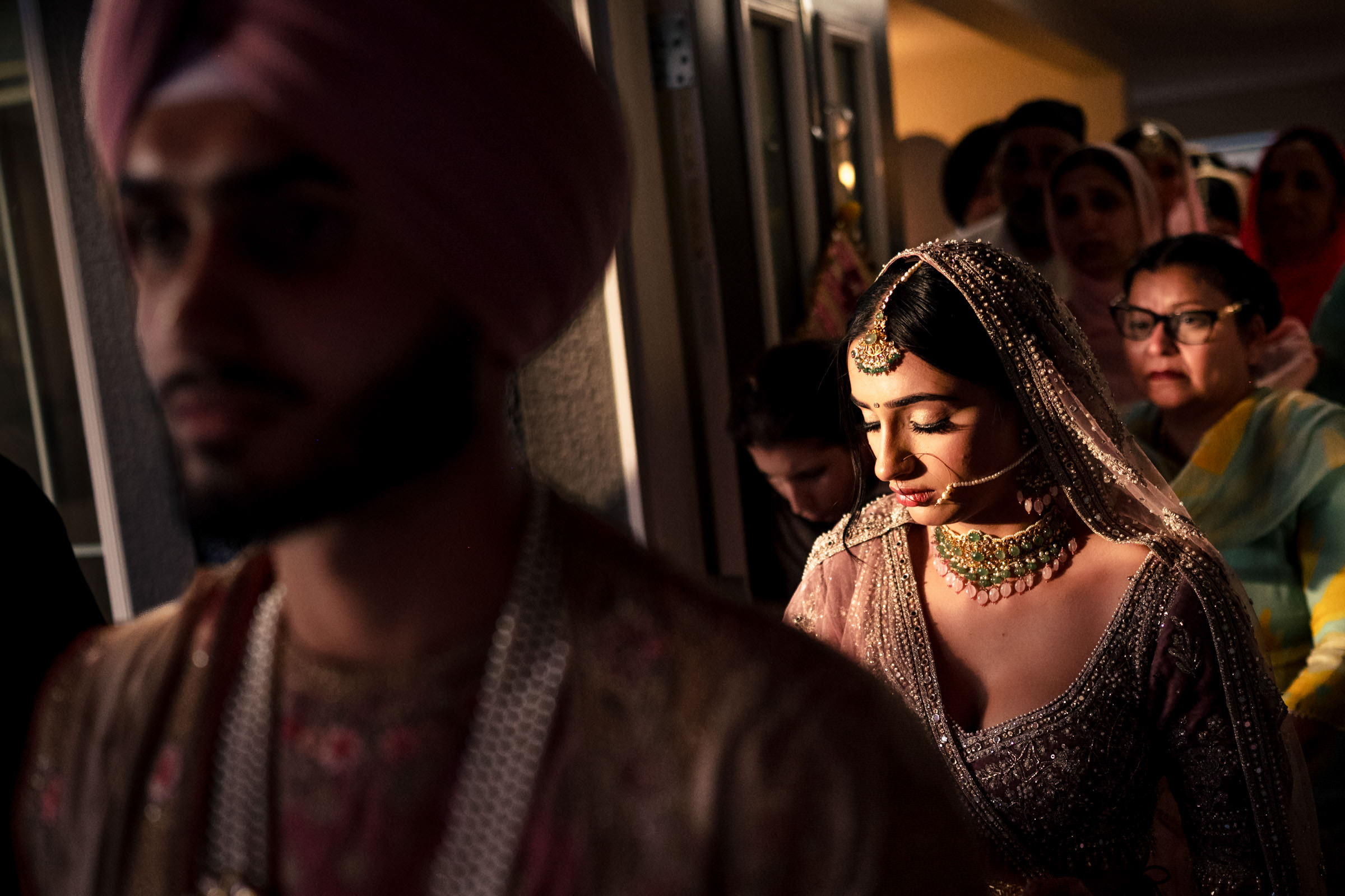 A bride in traditional attire with a jeweled necklace and headpiece graces one of our favourite wedding photos, walking behind a groom in a turban. The scene captures the essence of the ceremony, with attendees dressed vibrantly in the background.