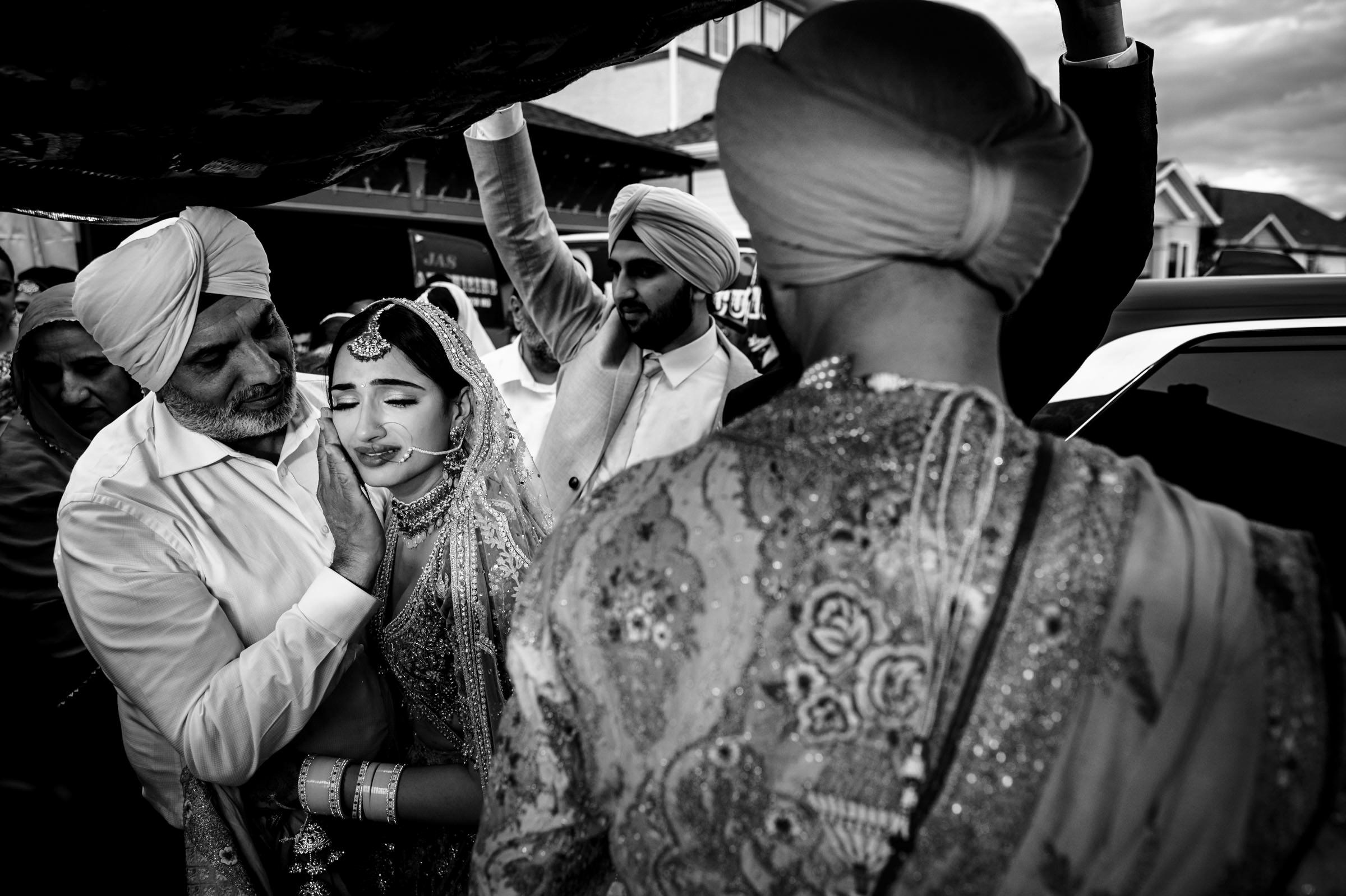 A black and white image captures one of the favourite wedding photos, highlighting an emotional moment at an Indian wedding. A bride in traditional attire is embraced by an older man under a decorated canopy, surrounded by others in traditional dress.