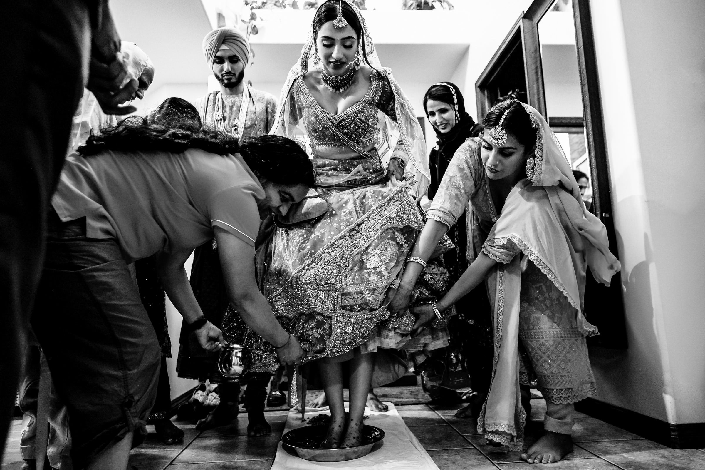 A bride in traditional attire steps into a bowl of liquid, surrounded by people assisting her. This cherished moment, often featured in favourite wedding photos, captures a cultural ritual with intricate patterns on the bride's outfit and attentive expressions on the faces around her.
