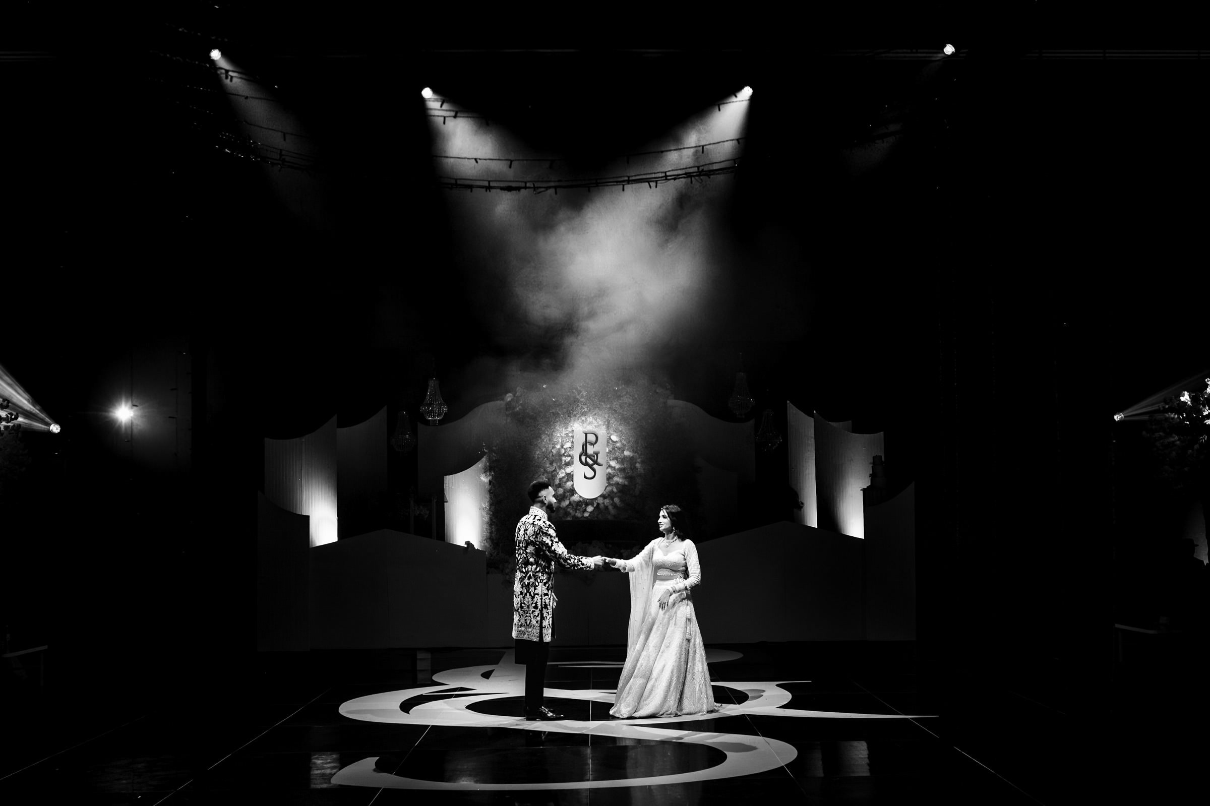 A black and white image captures one of their favourite wedding photos: a couple holding hands on a stage. The spotlight illuminates them, creating a dramatic atmosphere. In the background, a large monogram featuring the letters "P" and "S" adds an elegant touch.