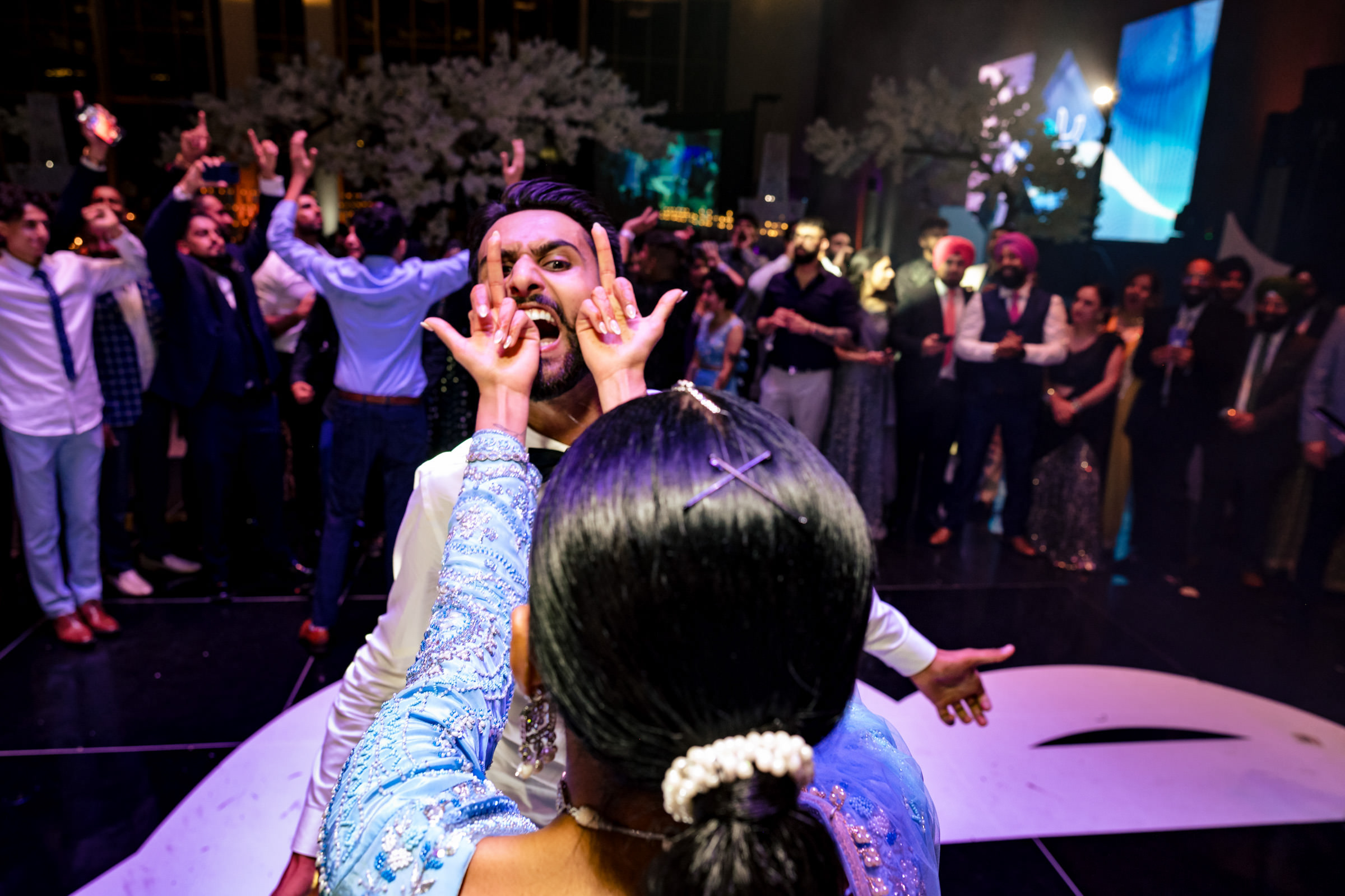 A lively dance floor scene unfolds as a woman playfully pinches a man's cheeks, capturing a moment likely to be among their favorite wedding photos. The man smiles broadly amid the formal-attired guests, with vibrant lighting and decorations setting the perfect celebratory backdrop.