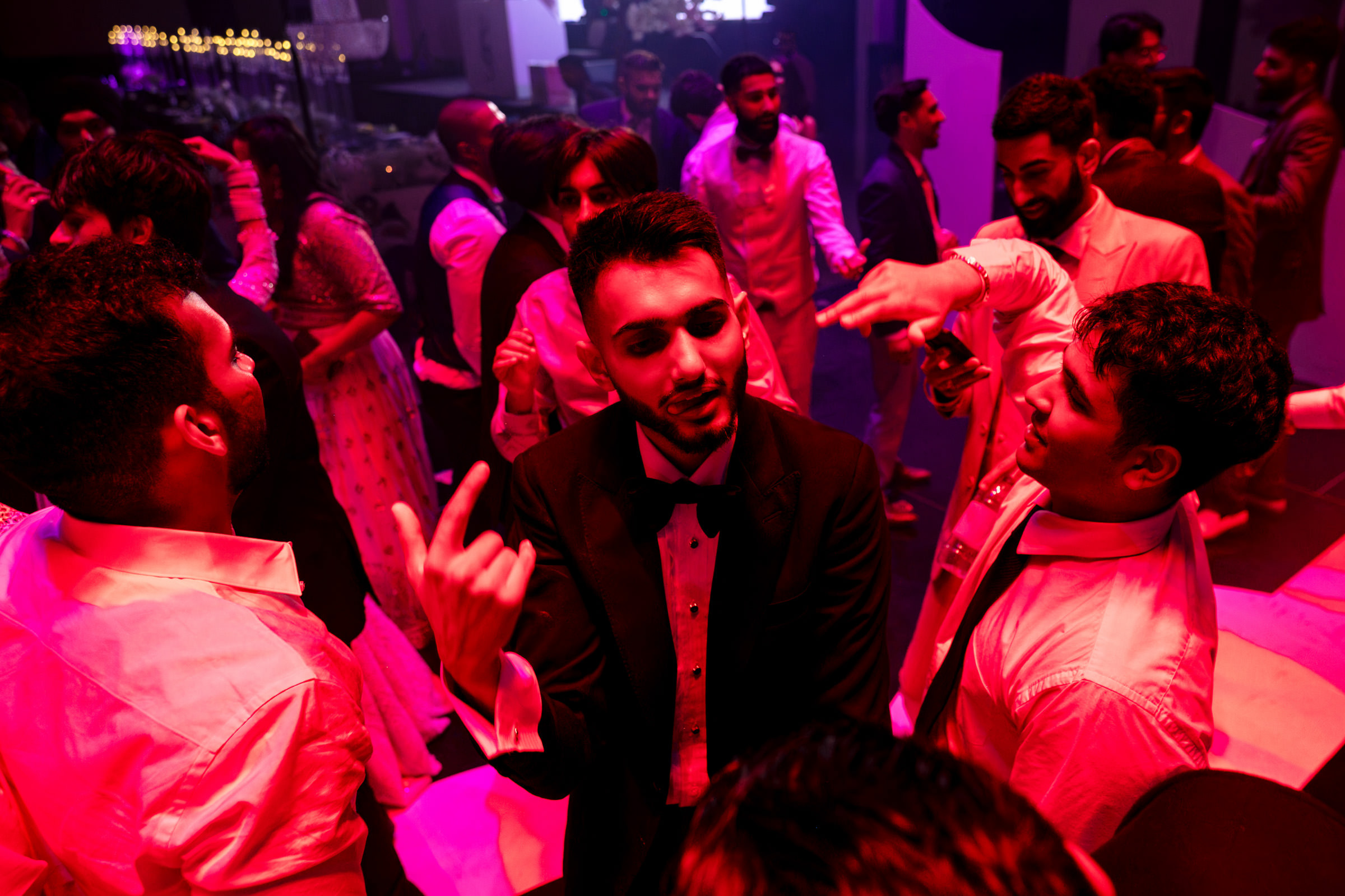 A lively party scene unfolds as a man in a tuxedo, featured in one of my favourite wedding photos, is surrounded by friends on the dance floor, all illuminated by red and purple lighting. In their formal attire, guests are joyfully chatting and dancing the night away.
