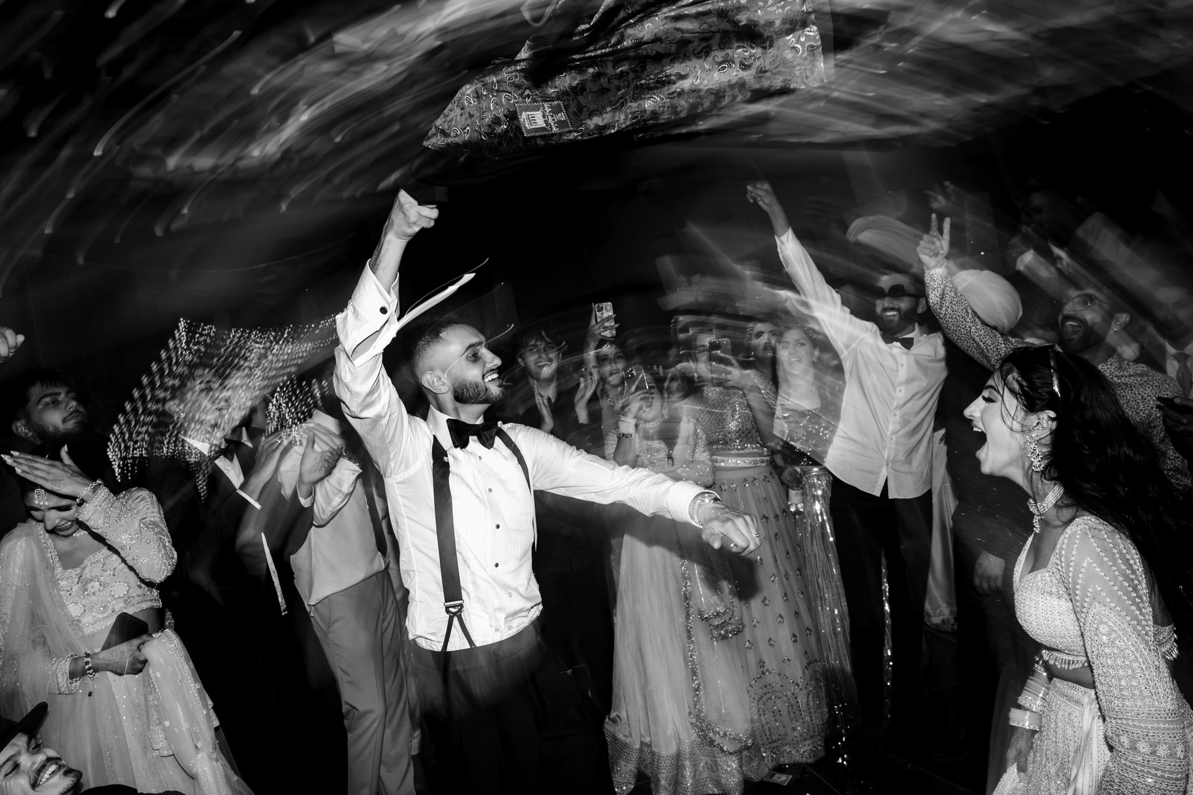 A black and white photo captures a lively scene at a party or wedding, with people dancing energetically. One of the favourite wedding photos shows a person in a hat and suspenders raising their arm enthusiastically, creating a dynamic, swirling effect in the image.
