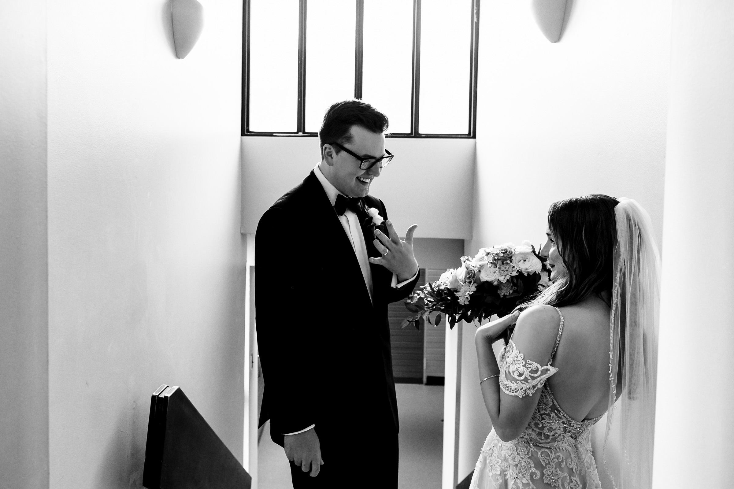 Black and white image of a couple on a staircase. The groom, in tuxedo and glasses, excitedly shows his wedding ring to the bride. The bride beams, holding a bouquet and wearing her veil. Natural light illuminates them in one of their favourite wedding photos.