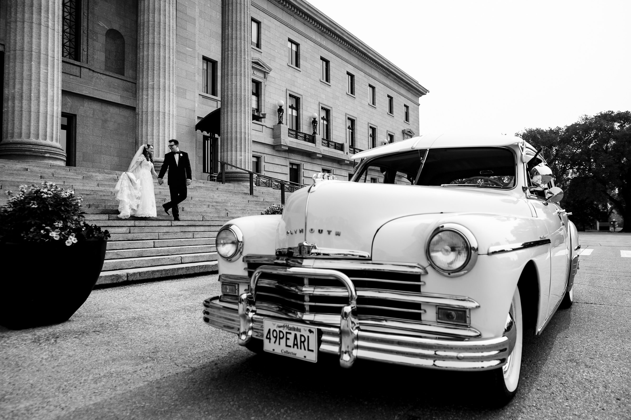 A bride and groom, featured in one of their favorite wedding photos, walk hand in hand down the steps of a grand building. In the foreground, a vintage white Plymouth car with a "49PEARL" license plate is parked. The timeless scene is captured in black and white.