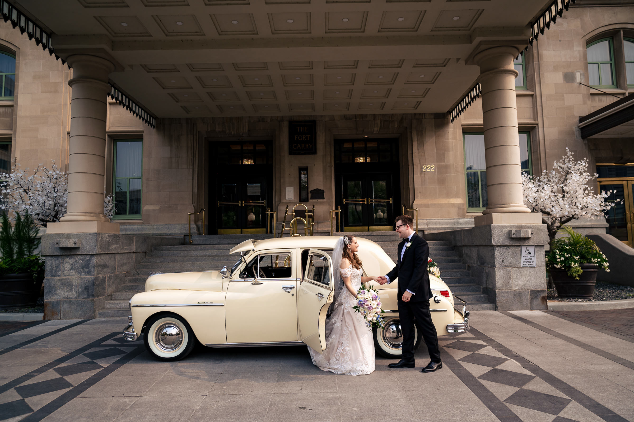 A bride and groom stand in front of a vintage cream car outside a grand building with columns, capturing one of their favourite wedding photos. The groom, in a black suit, assists the bride, in a white gown, as she exits the car holding a colorful bouquet. The setting is formal and elegant.