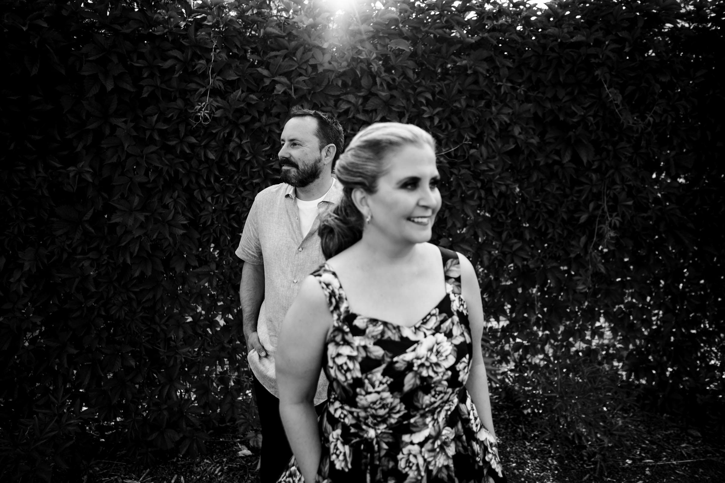 A black and white photo captures a couple outdoors in what is surely one of their favourite wedding photos. The woman, in a floral dress, smiles brightly while the man stands behind with his hands in his pockets. A leafy backdrop and sunlight enhance the scene.