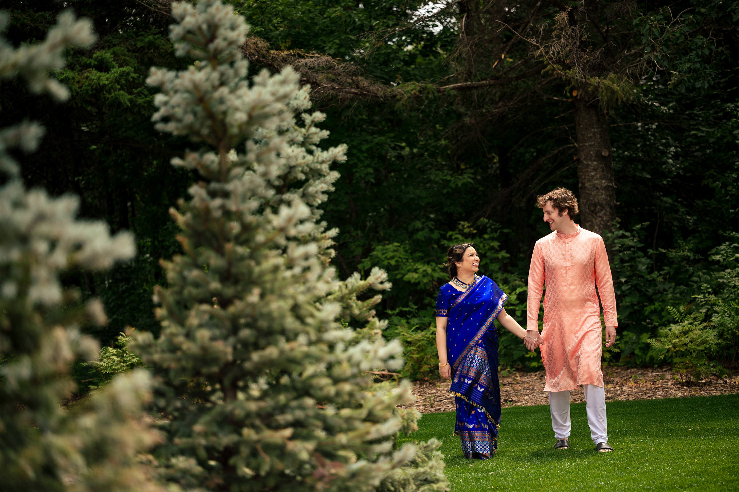 A couple holding hands, smiling at each other as they stroll through a garden. The woman in a blue sari and the man in a peach kurta with white pants are enveloped by evergreen trees and lush greenery—a scene reminiscent of their favorite wedding photos.