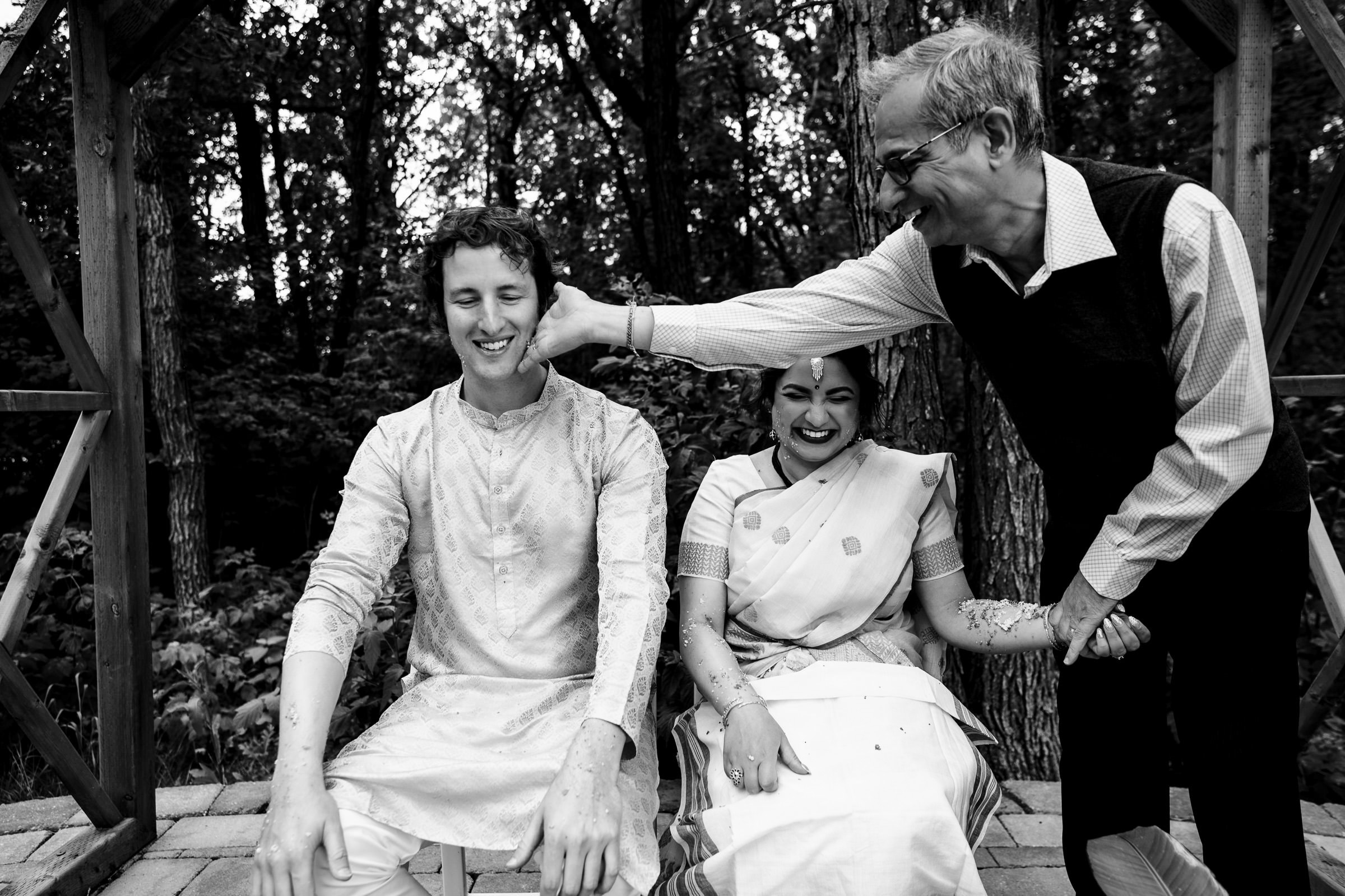 A black and white photo, one of our favorite wedding photos, captures two seated individuals in traditional attire. They smile as an older man playfully pinches the younger man's cheek. The scene is set outdoors with trees in the background.