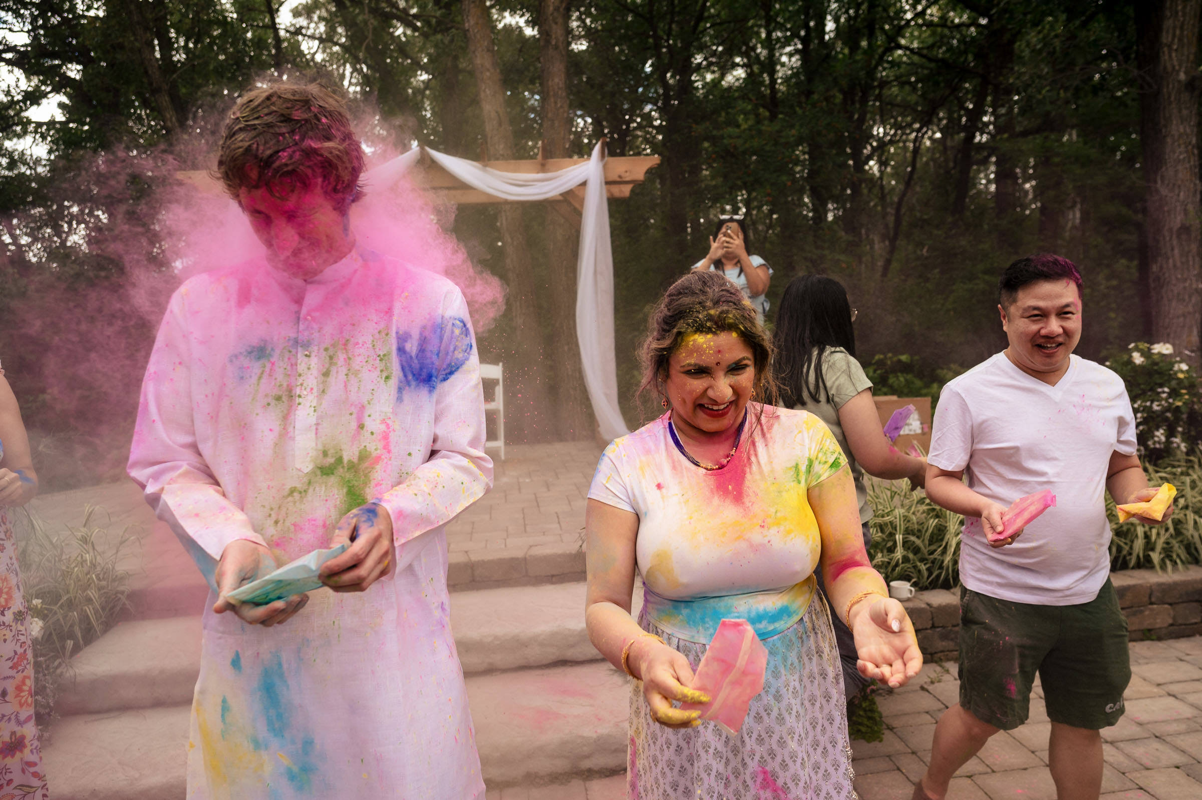 People enjoying a colorful Holi celebration outdoors, reminiscent of their favorite wedding photos. They are covered in vibrant powders and smiling, with trees and a decorated archway visible in the background.