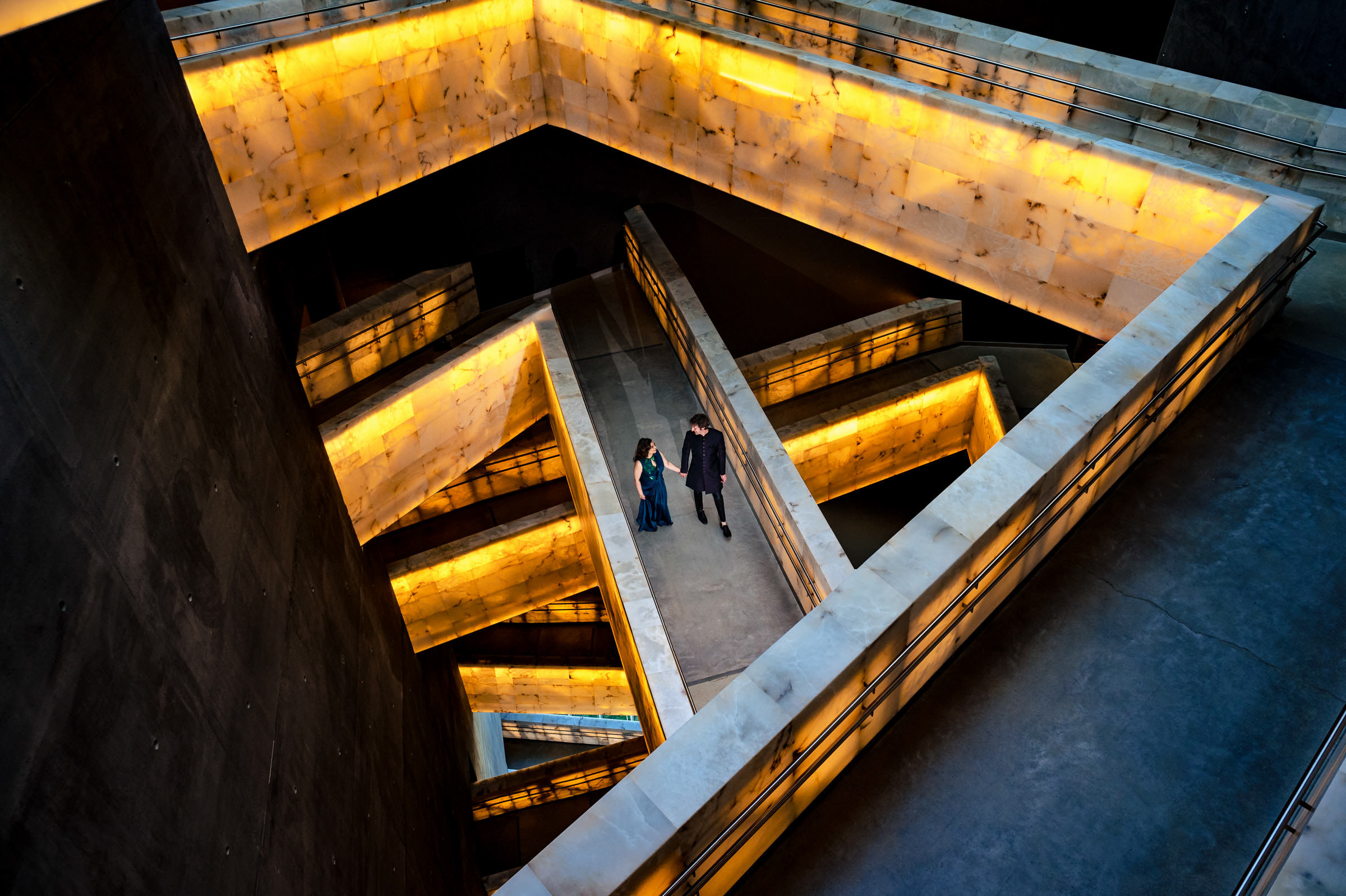 The couple descends a series of illuminated, angular staircases with glowing yellow lights, capturing one of their favourite wedding photos. The zigzag pattern in the modern architectural space creates a warm, geometric atmosphere.