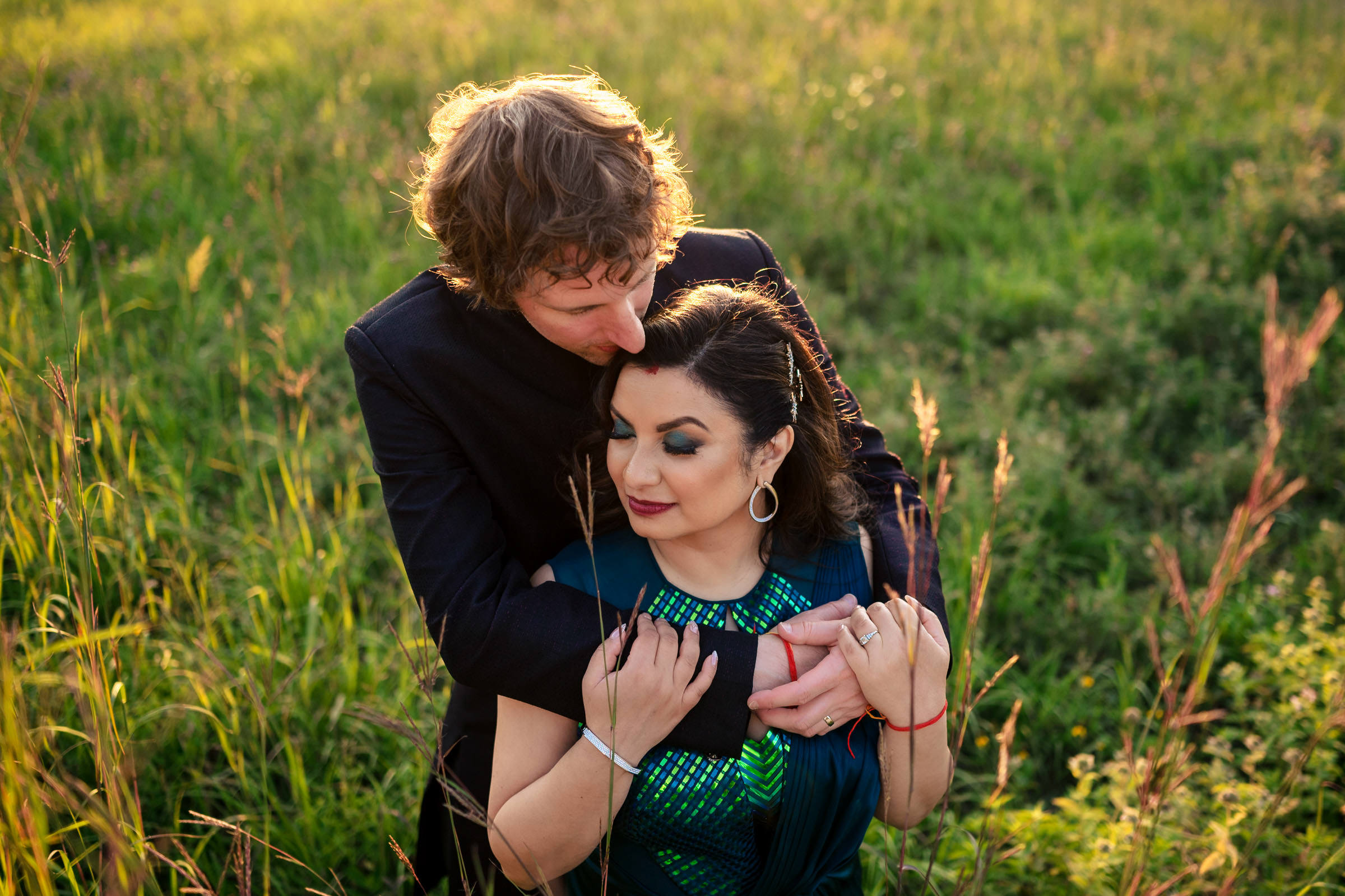 In one of their favourite wedding photos, a couple embraces in a sunlit field. The man, with curly hair, gently holds and kisses the woman's head. The woman, with long dark hair and wearing a green dress, looks content. Tall grass surrounds them under a clear sky.