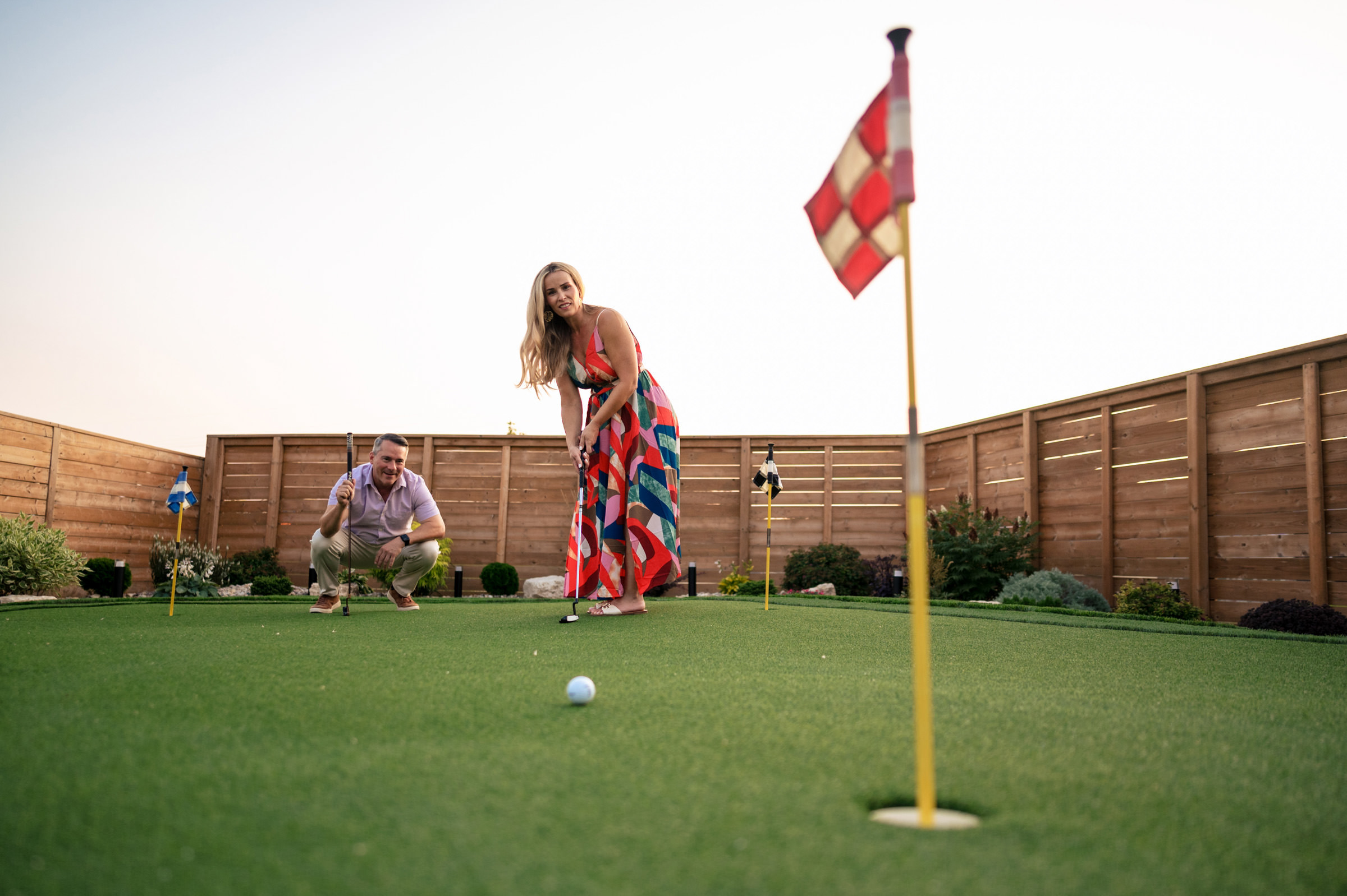 A woman in a colorful dress is putting a golf ball on a small green, reminiscent of favourite wedding photos. Nearby, a man crouches, observing closely. Wooden fencing and plants surround the scene under a clear sky with red and white flags fluttering gently.