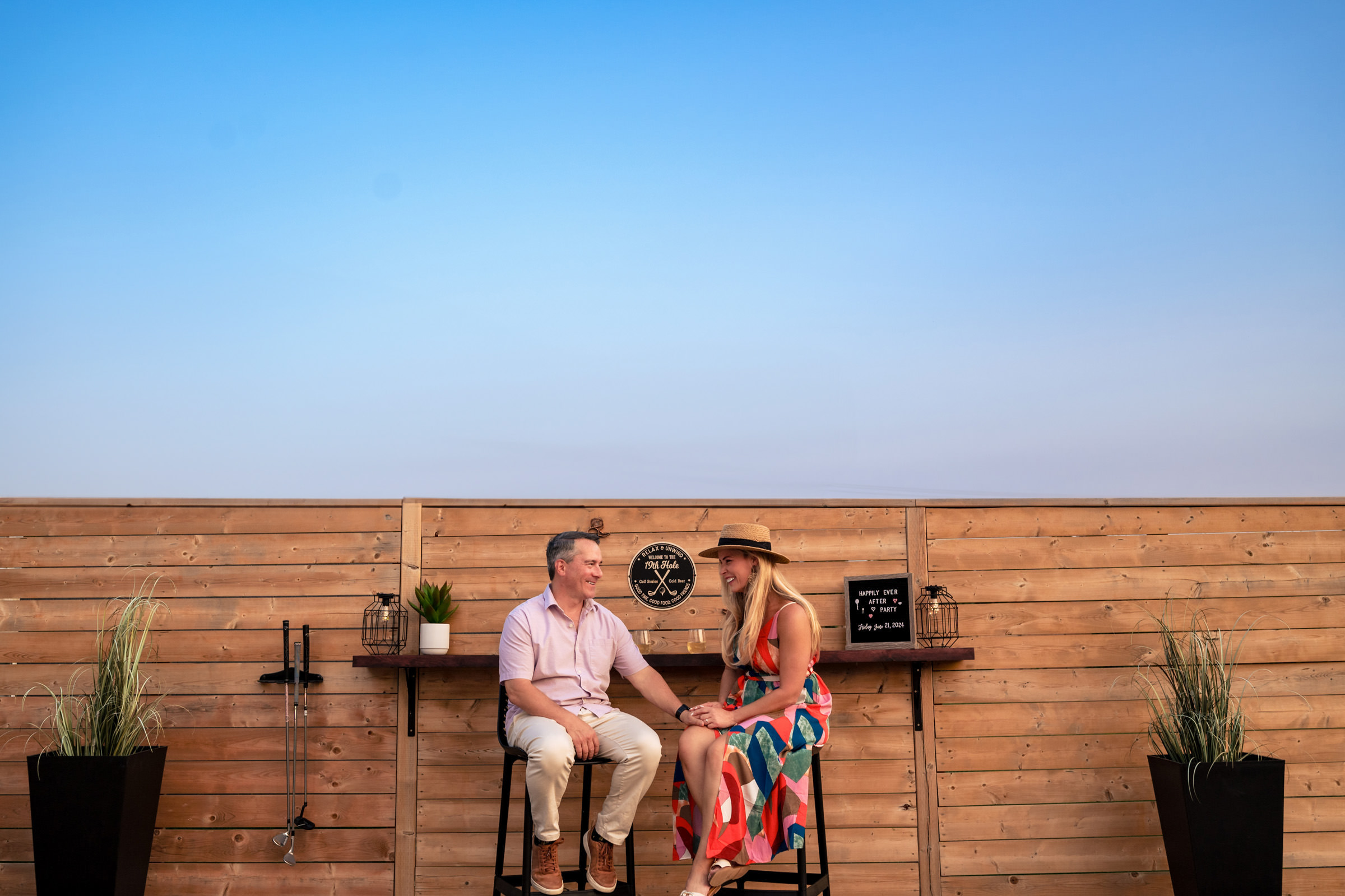 A man and woman sit on stools in front of a wooden fence under a clear sky, reminiscent of their favourite wedding photos. The man wears a light shirt and pants, while the woman dons a colorful dress and hat. They seem engrossed in conversation, surrounded by potted plants and small decorative items.