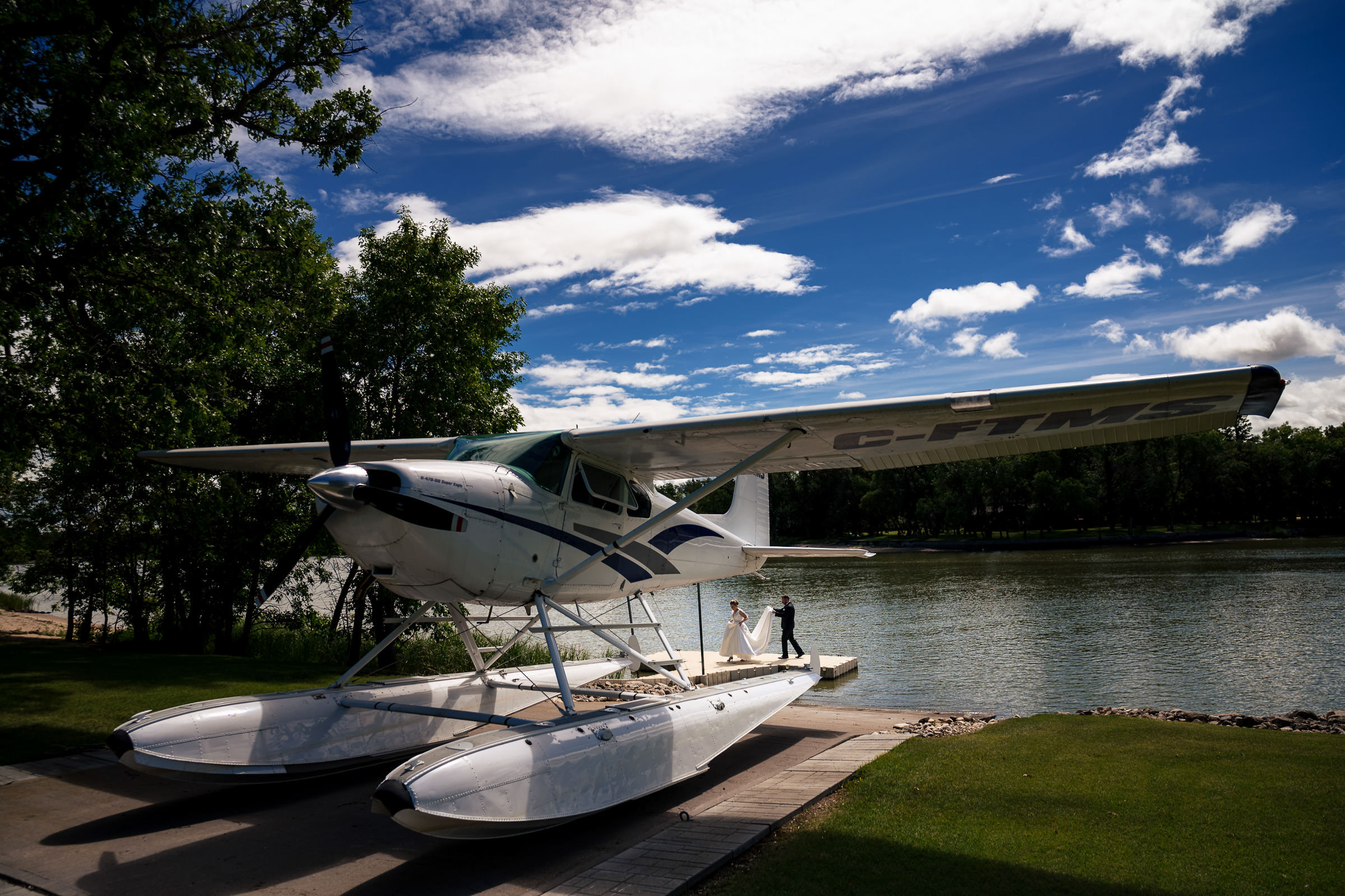 A white floatplane is parked by the water on a sunny day, with a few people standing nearby. The backdrop of trees and a bright blue sky with scattered clouds makes it the perfect setting for capturing your favorite wedding photos.