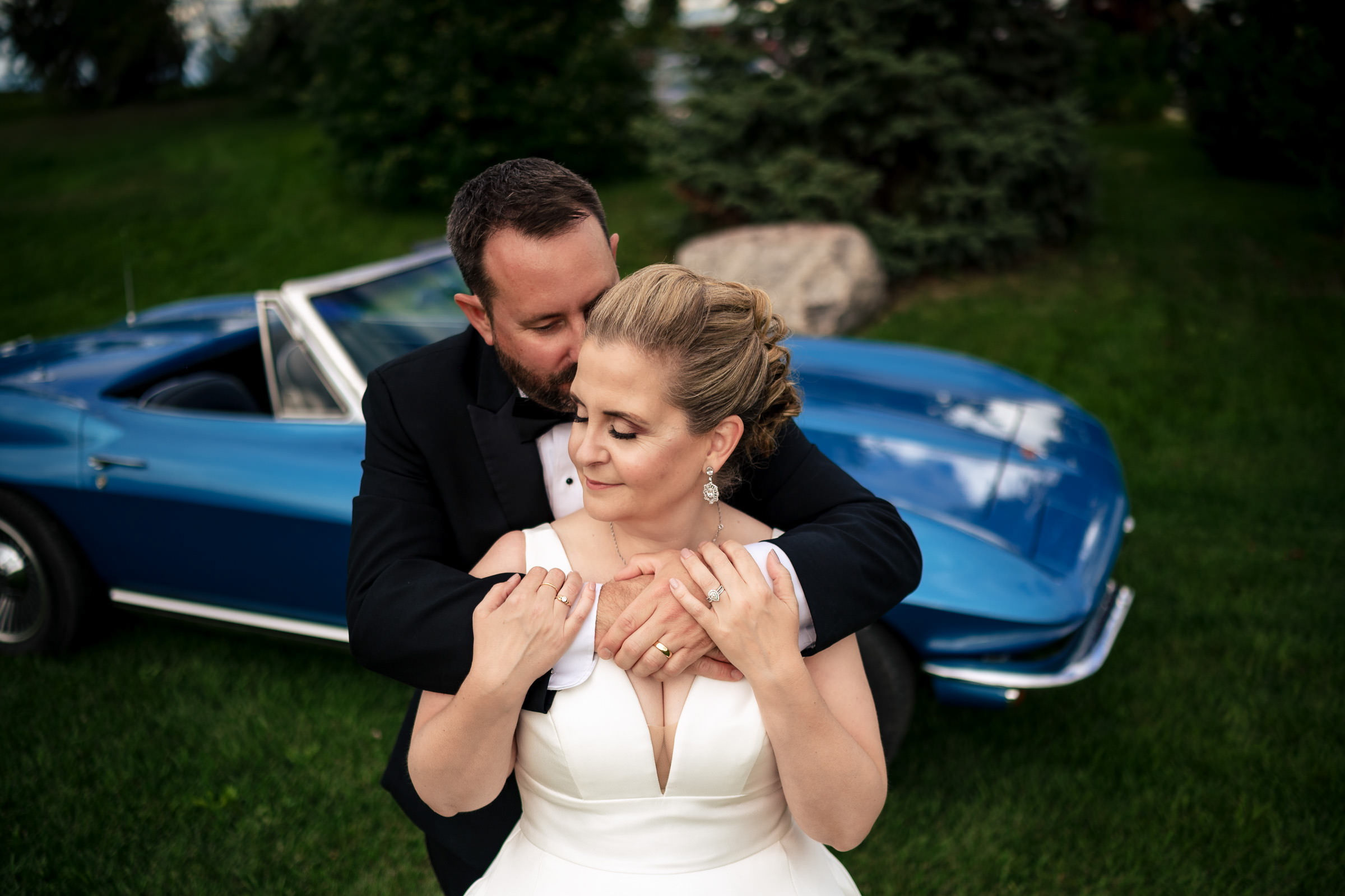 A couple embraces in front of a blue vintage convertible on the grassy area, captured among their favorite wedding photos. The woman wears a white dress, the man a black suit. Both have closed eyes, and the scene is serene and romantic.