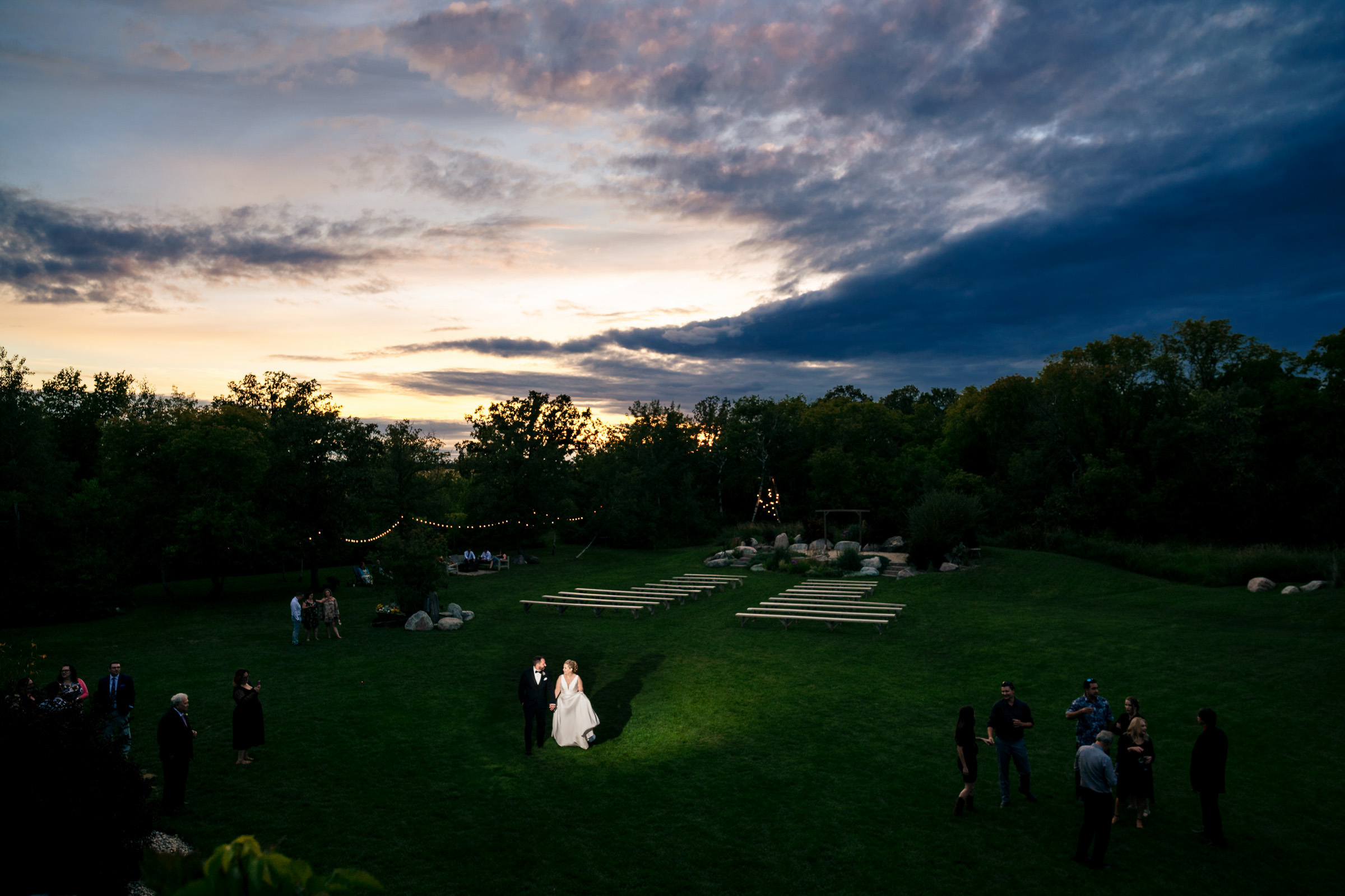 A couple getting married in an open field at dusk creates one of those favourite wedding photos. They stand under a spotlight surrounded by guests, with a dramatic evening sky in the background. String lights and empty wooden benches enhance the enchanting atmosphere.