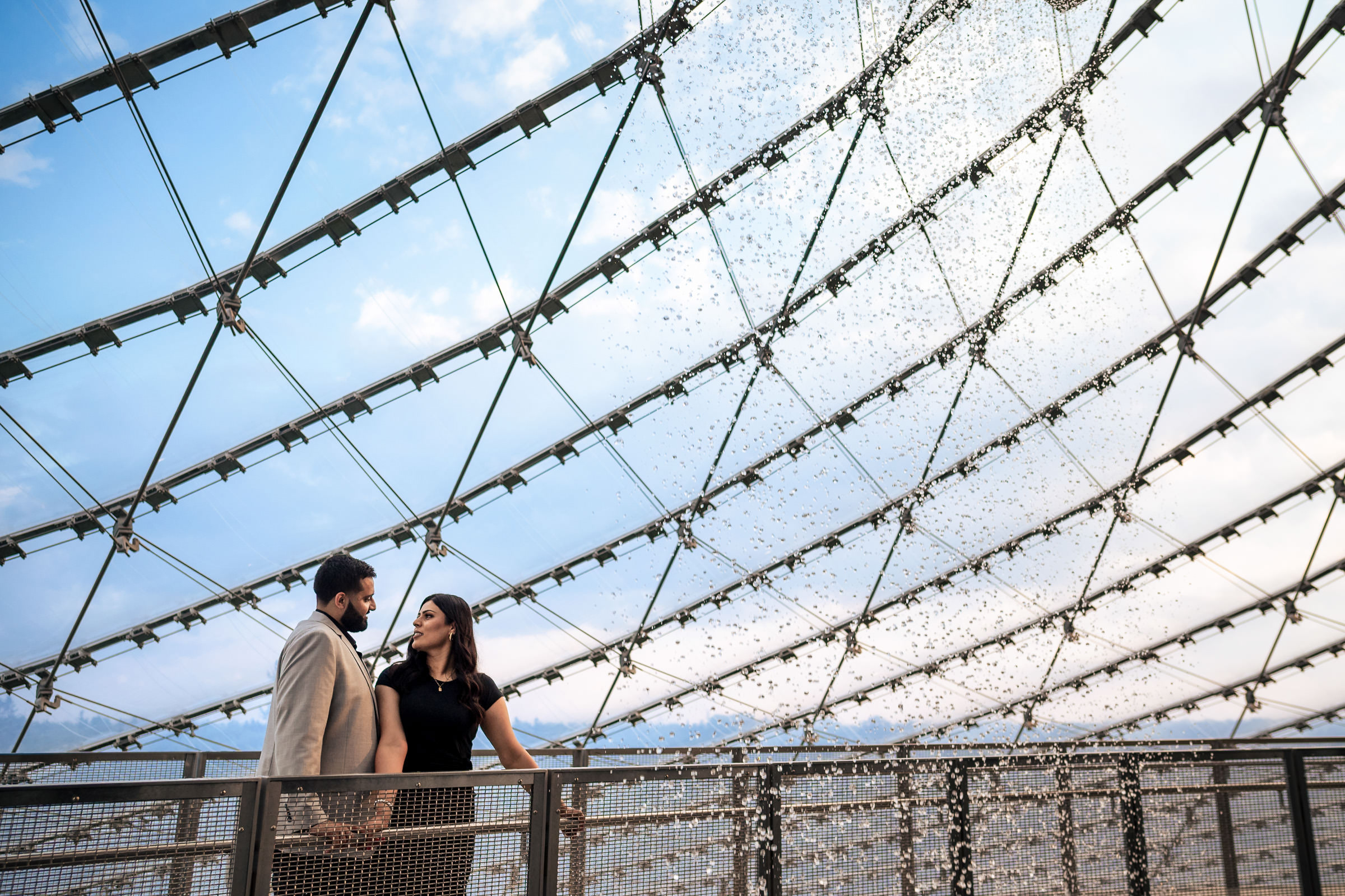 A couple holds hands on a modern glass walkway with a geometric metal lattice overhead, set against a blue sky. They appear content and engaged in conversation, capturing one of their favourite wedding photos.