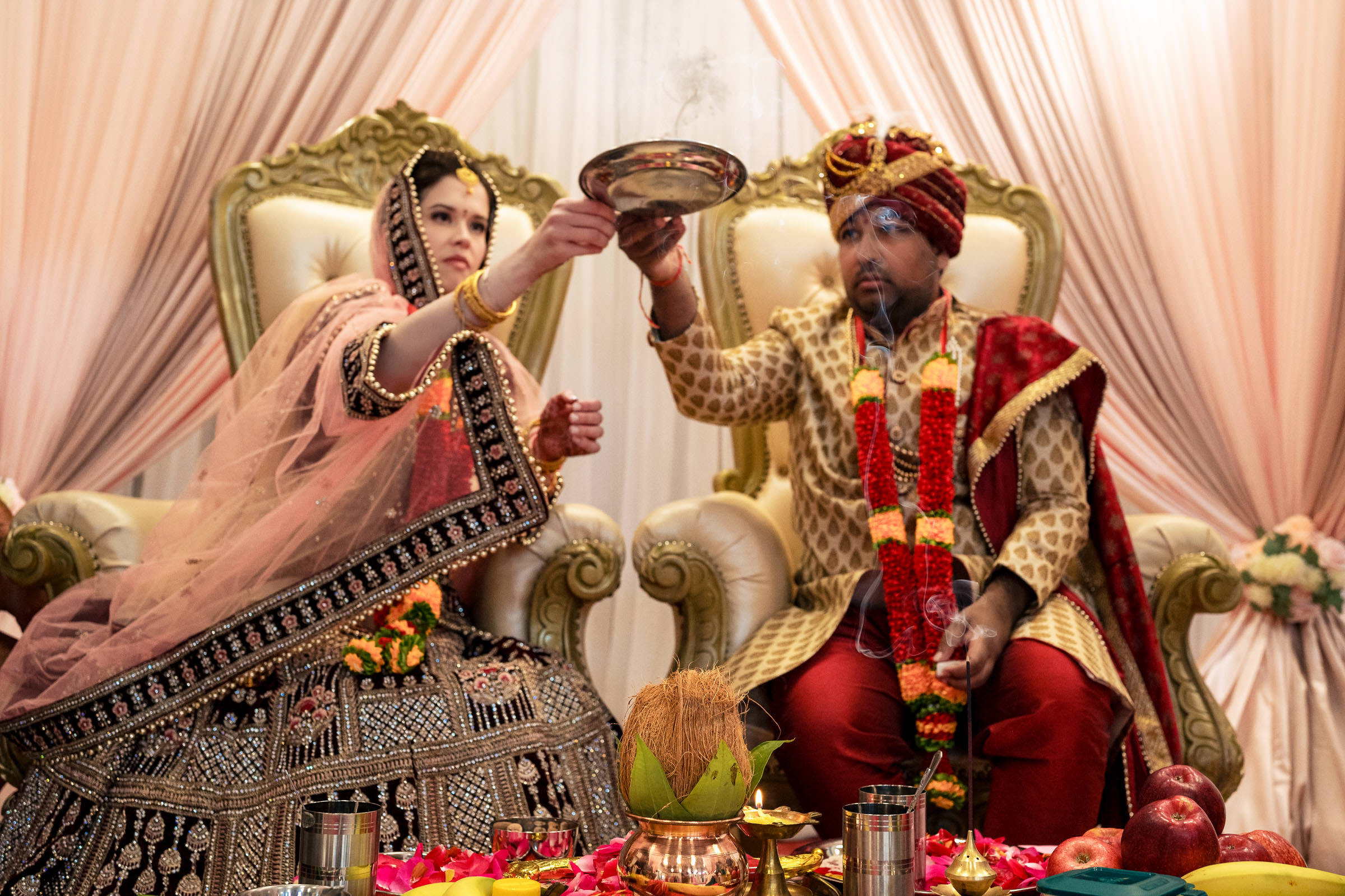 A couple in traditional Indian attire poses for favorite wedding photos during a cultural ceremony. Seated on ornate chairs and surrounded by ritual items, the woman dazzles in a detailed lehenga while the man dons a sherwani and turban, both lifting a tray together.