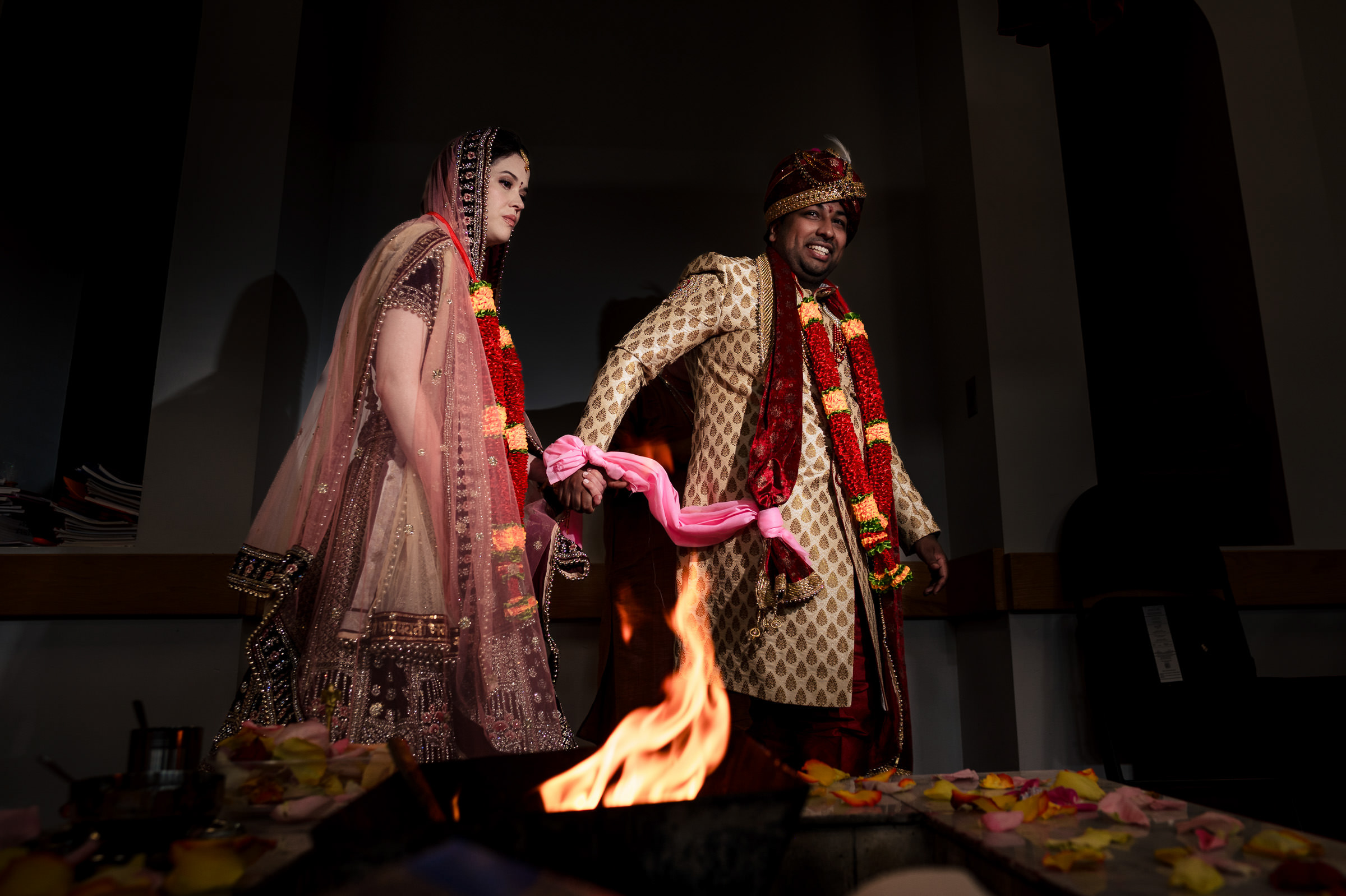 A couple in traditional wedding attire walks around a sacred fire, holding hands and performing a ritual. The bride wears a pink lehenga and the groom, a golden sherwani, both adorned with floral garlands. Multicolored petals surround the fire, capturing one of their favorite wedding photos.