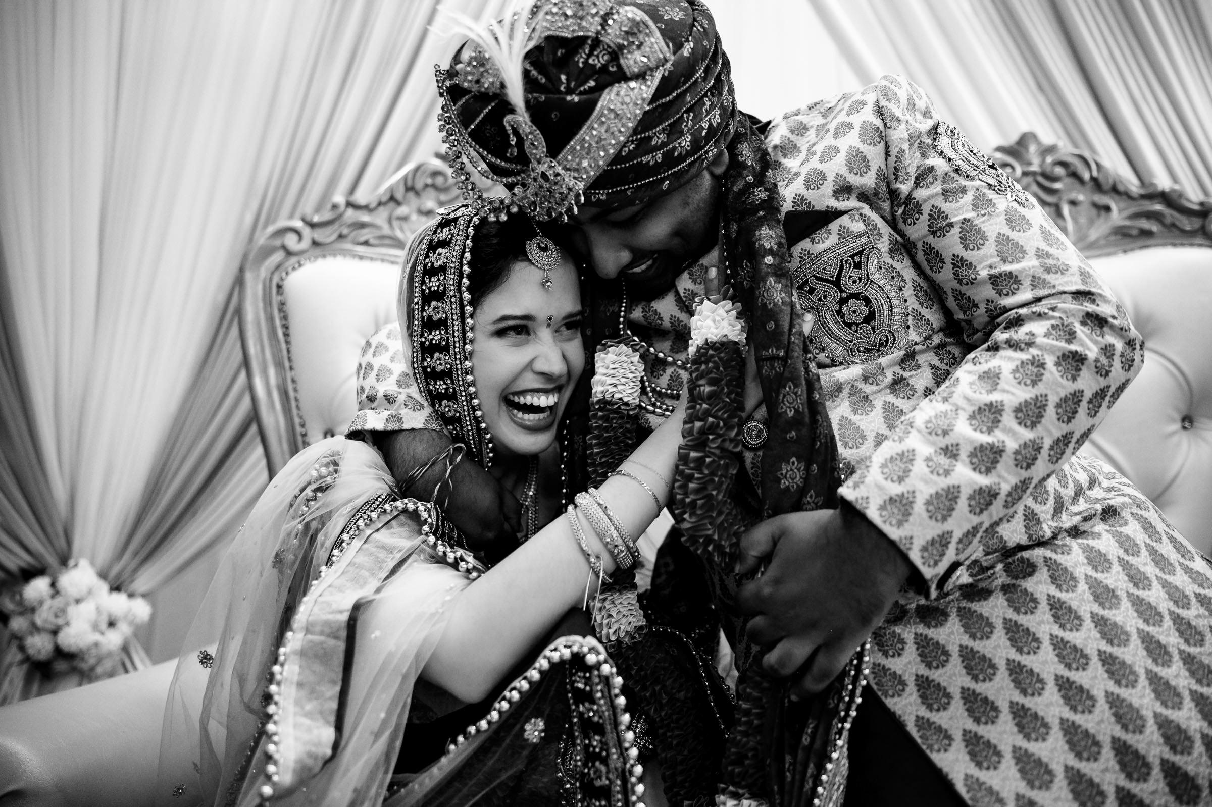 A joyful couple, captured in one of their favorite wedding photos, dressed in traditional Indian attire, share a moment of laughter. The bride dazzles in a detailed saree and ornate jewelry, while the groom stuns in a patterned sherwani and turban as they sit closely on an ornate couch.