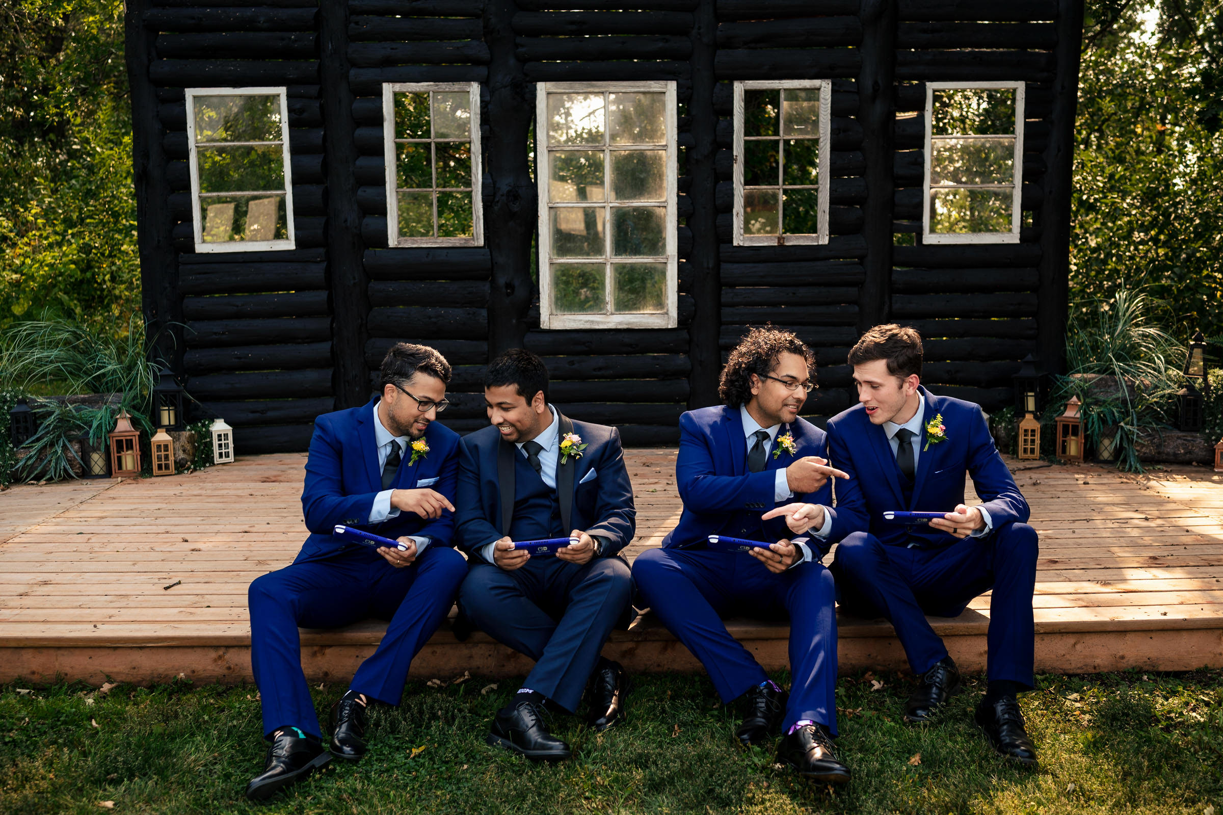 Four men in blue suits sit on a wooden deck in front of a rustic cabin, each holding a sheet of paper like they're reviewing their favourite wedding photos. They smile and point at each other's papers, surrounded by greenery and decorative lanterns.