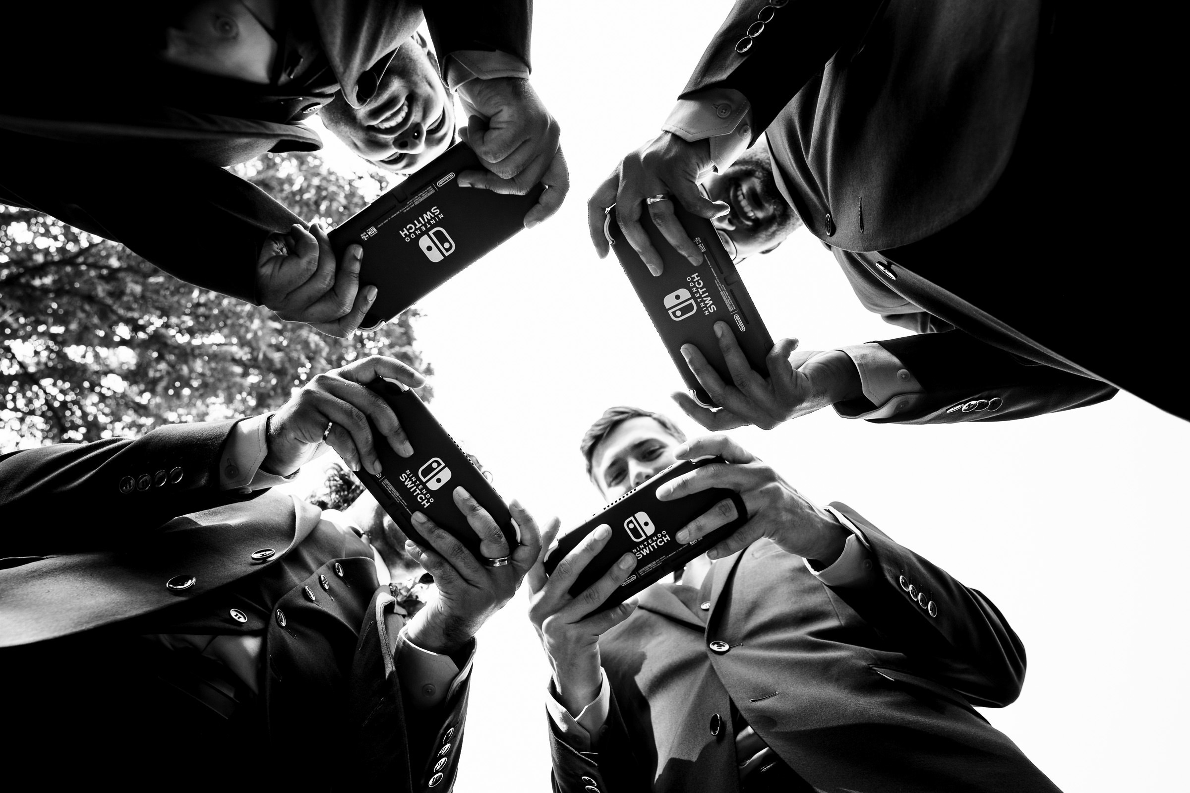 Five people in suits stand in a circle, viewed from below, each engrossed in portable gaming consoles. The black-and-white image captures their engaged expressions and the symmetry of their gathering, reminiscent of favourite wedding photos with its unique charm and focused connection.