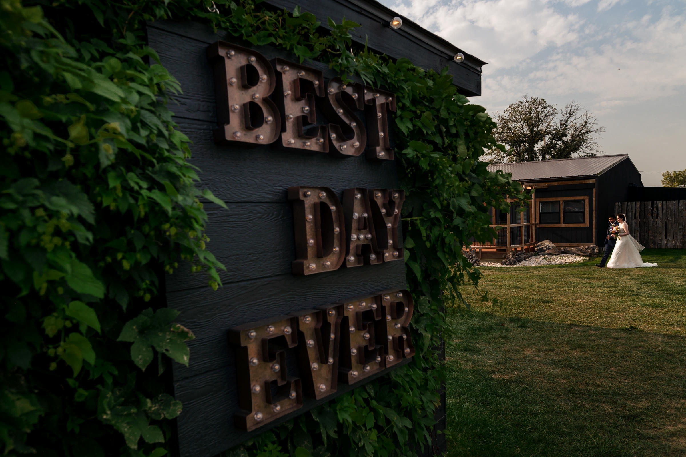 A large sign reading "BEST DAY EVER" is adorned with vines on a dark wall, perfectly framing one of their favourite wedding photos. In the background, a bride and groom stand together on a lawn in front of a rustic building under a partly cloudy sky.