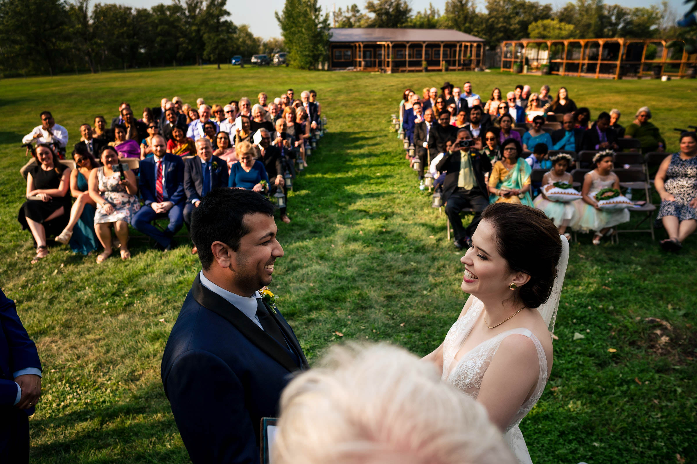 A bride and groom stand facing each other, smiling, during an outdoor ceremony that would soon become one of their favorite wedding photos. Behind them, guests are seated in rows on a grassy lawn under a clear sky, with trees and a wooden building adding charm to the background.
