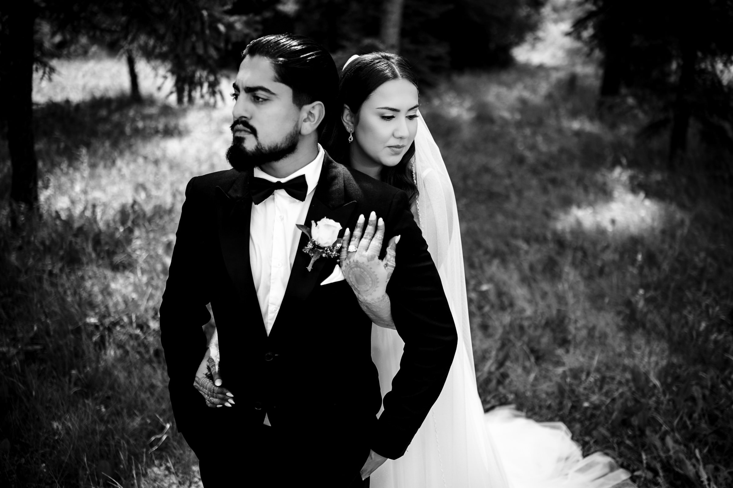 A black and white photo captures one of their favourite wedding moments: a groom in a tuxedo and bow tie stands thoughtfully in a grassy, wooded area while the bride, in veil and gown, lovingly embraces him from behind as they both gaze into the distance.
