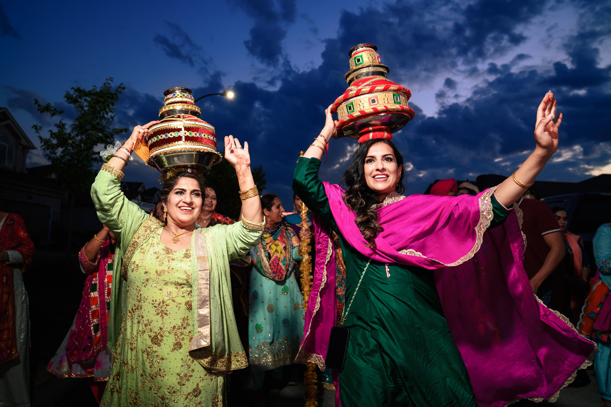 Two women in vibrant traditional attire joyfully dance with decorated pots on their heads at dusk, capturing the essence of cultural festivity against a dramatic evening sky. Their colorful clothing and joyous expressions are reminiscent of favourite wedding photos cherished for their lively spirit.