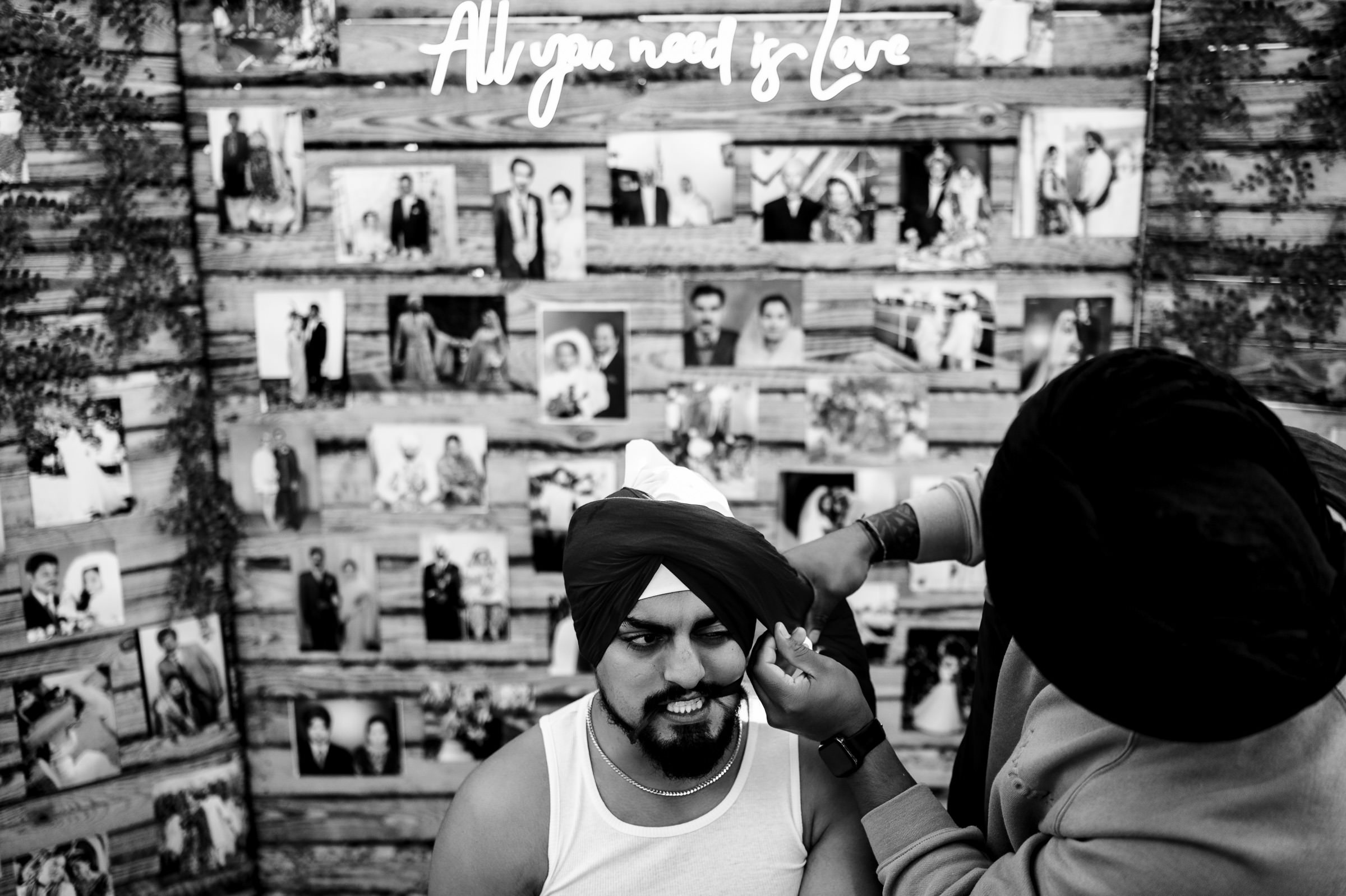 A person is adjusting the turban of another sitting on a chair, surrounded by favourite wedding photos. Above them, a neon sign glows, "All you need is love." The entire scene unfolds in black and white.