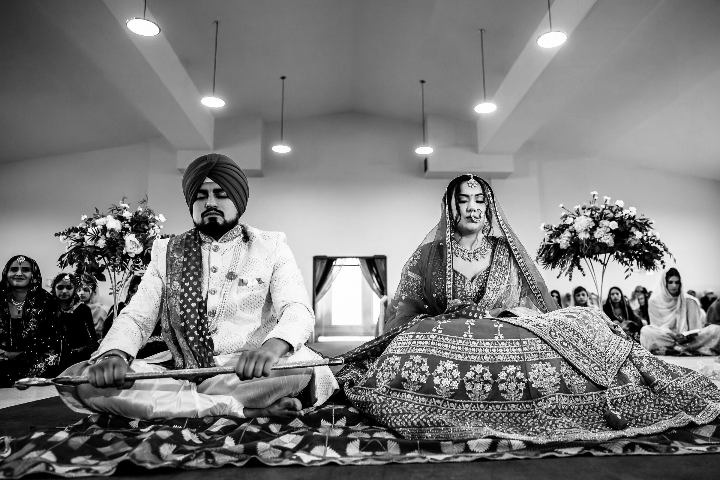 A bride and groom in traditional attire participate in a wedding ceremony indoors. The groom holds a staff, and the bride sits beside him in what could easily become one of their favourite wedding photos. Floral arrangements and guests adorn the background. The image is in black and white.