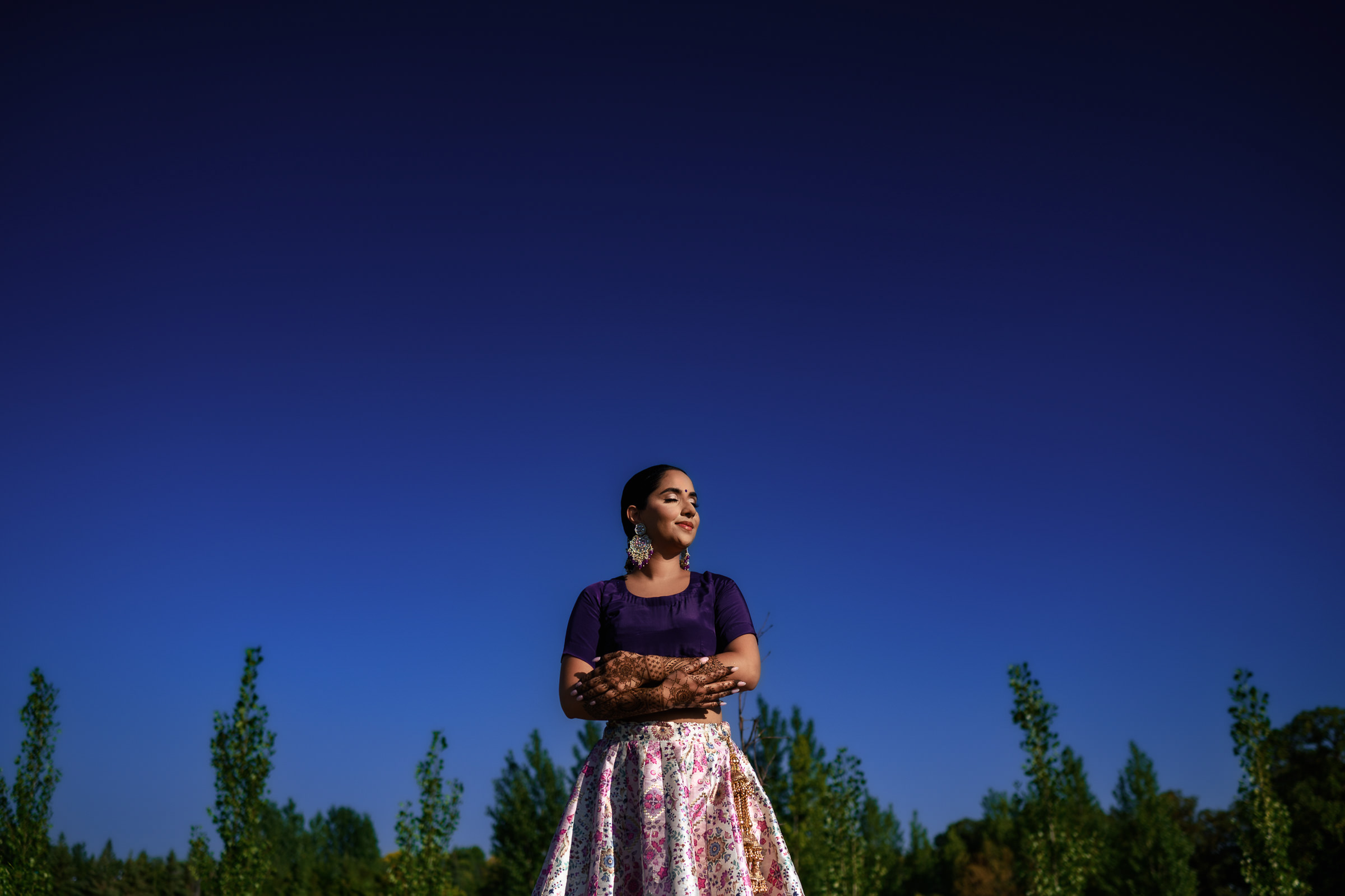 A woman stands confidently with her arms crossed against a backdrop of a vibrant blue sky and green trees. She wears a purple top and a floral skirt, reminiscent of favourite wedding photos, with her hair styled up and large earrings adding elegance to her look.
