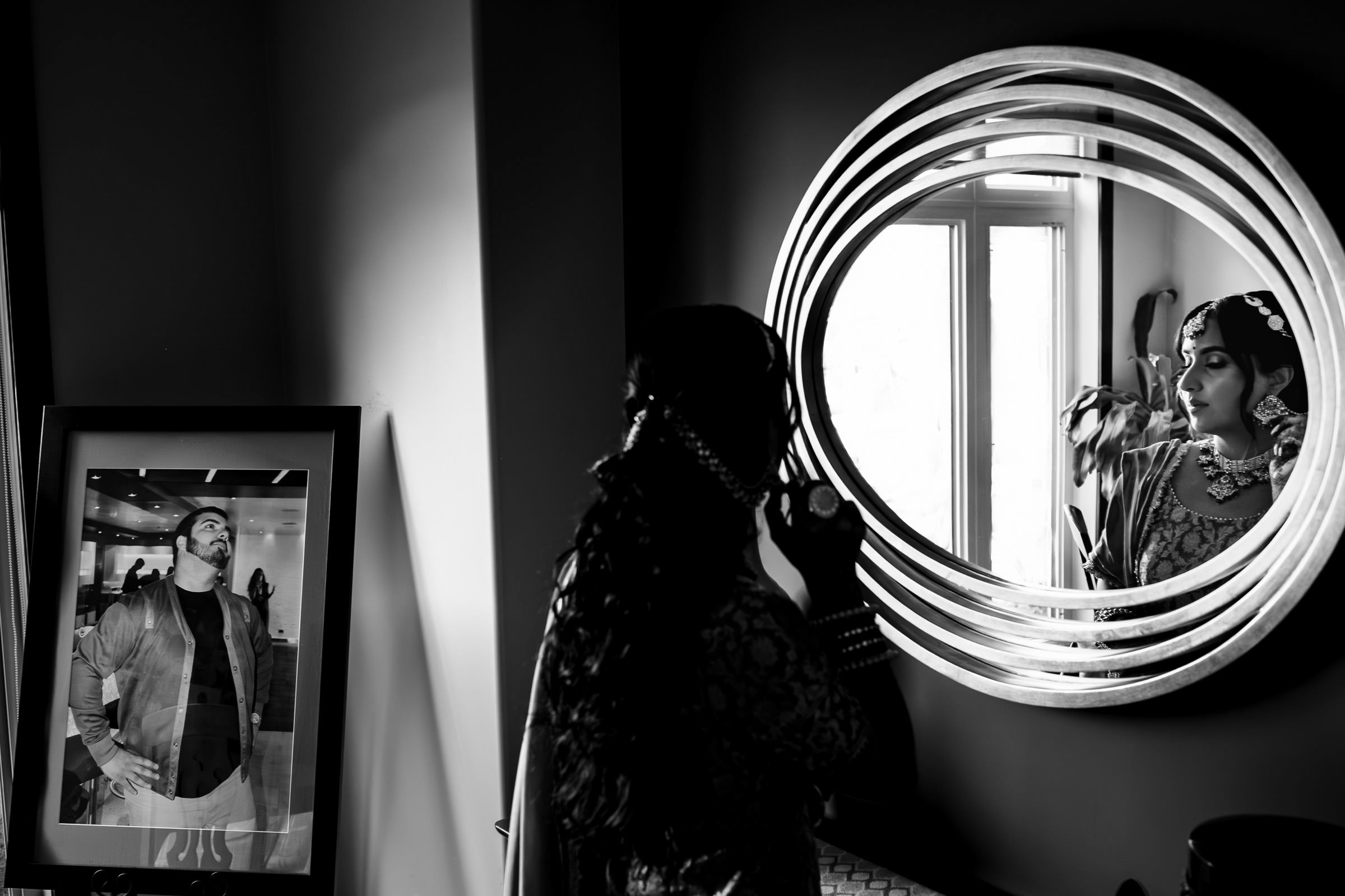 A woman in traditional attire applies makeup, gazing into a round mirror. Her reflection blends seamlessly with the room’s window. A framed photo of a man, reminiscent of favourite wedding photos, sits on the left. The image is elegantly captured in black and white.