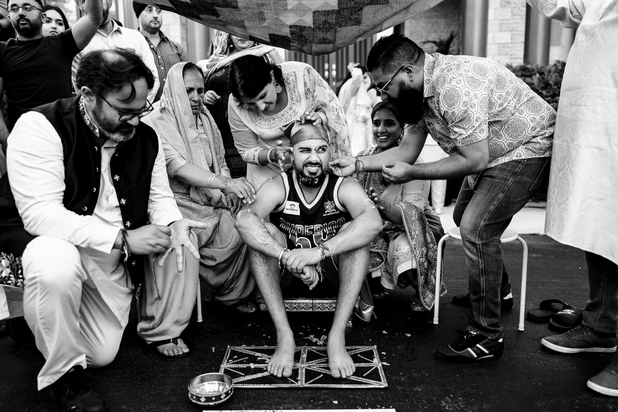A groom in a basketball jersey sits on a street, surrounded by joyful friends and family performing a ritual. They're pouring liquid and applying substance on him. A patterned cloth is held overhead as everyone laughs and celebrates. It's one of those favourite wedding photos captured in black and white.