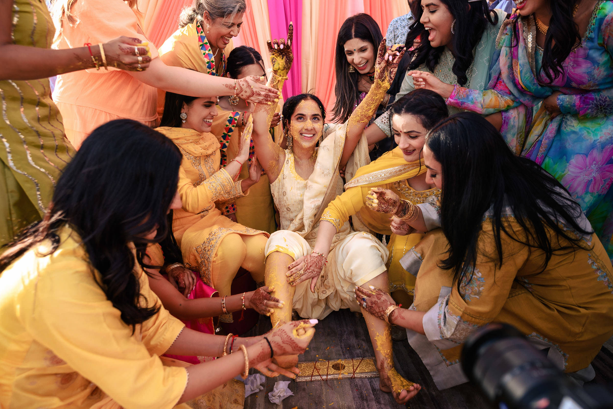 A joyful bride in her white and yellow outfit is surrounded by friends applying turmeric paste during a traditional pre-wedding ceremony. Everyone, dressed in colorful attire with pink and orange drapes around them, is all smiles. It's one of their favorite wedding photo moments.