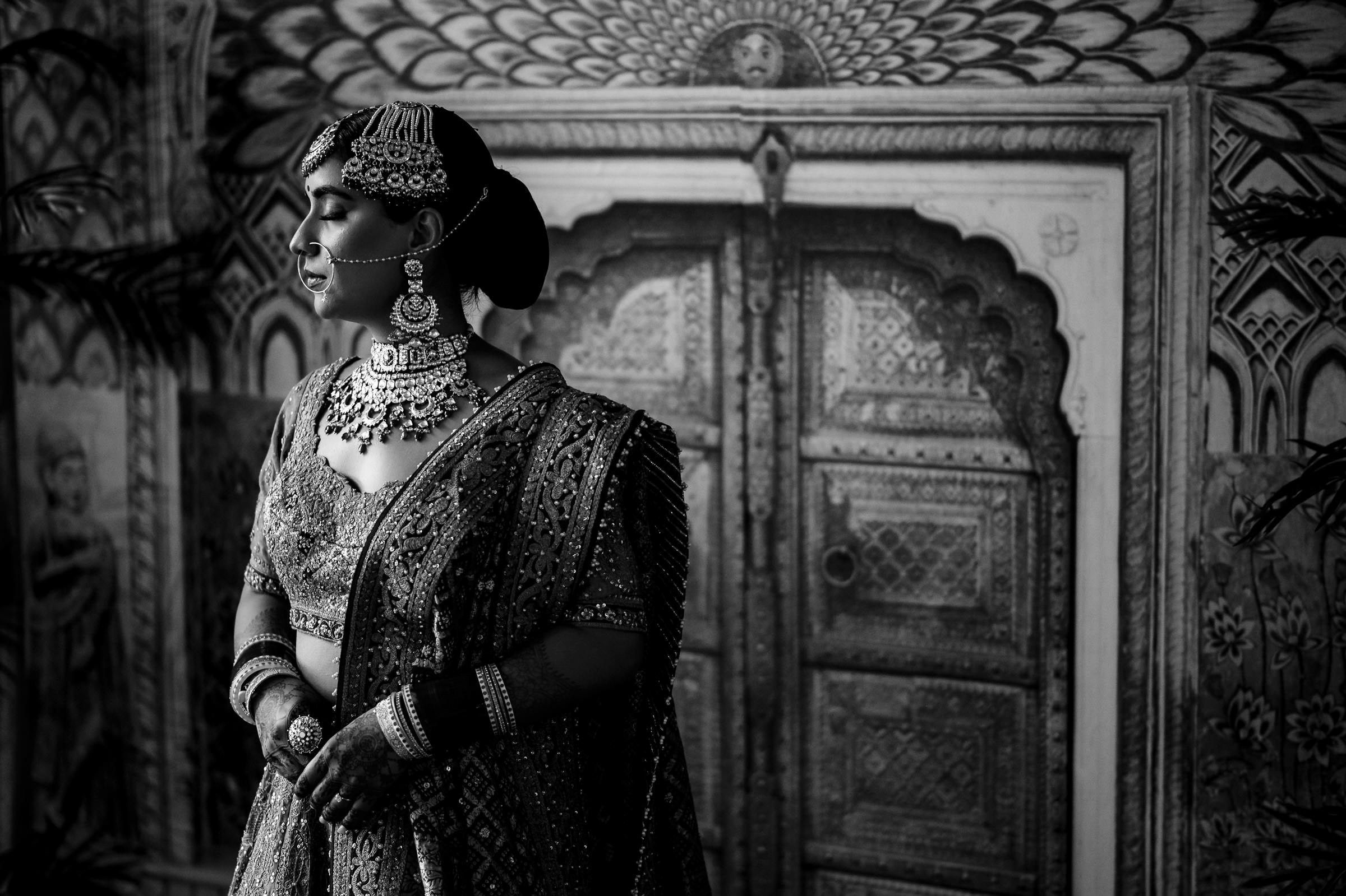 A woman in traditional Indian bridal attire stands gracefully before an intricately carved wooden door, capturing one of her favorite wedding photos. She wears a heavily embroidered lehenga, ornate jewelry, and a maang tikka, glancing to the side against the backdrop's detailed patterns.