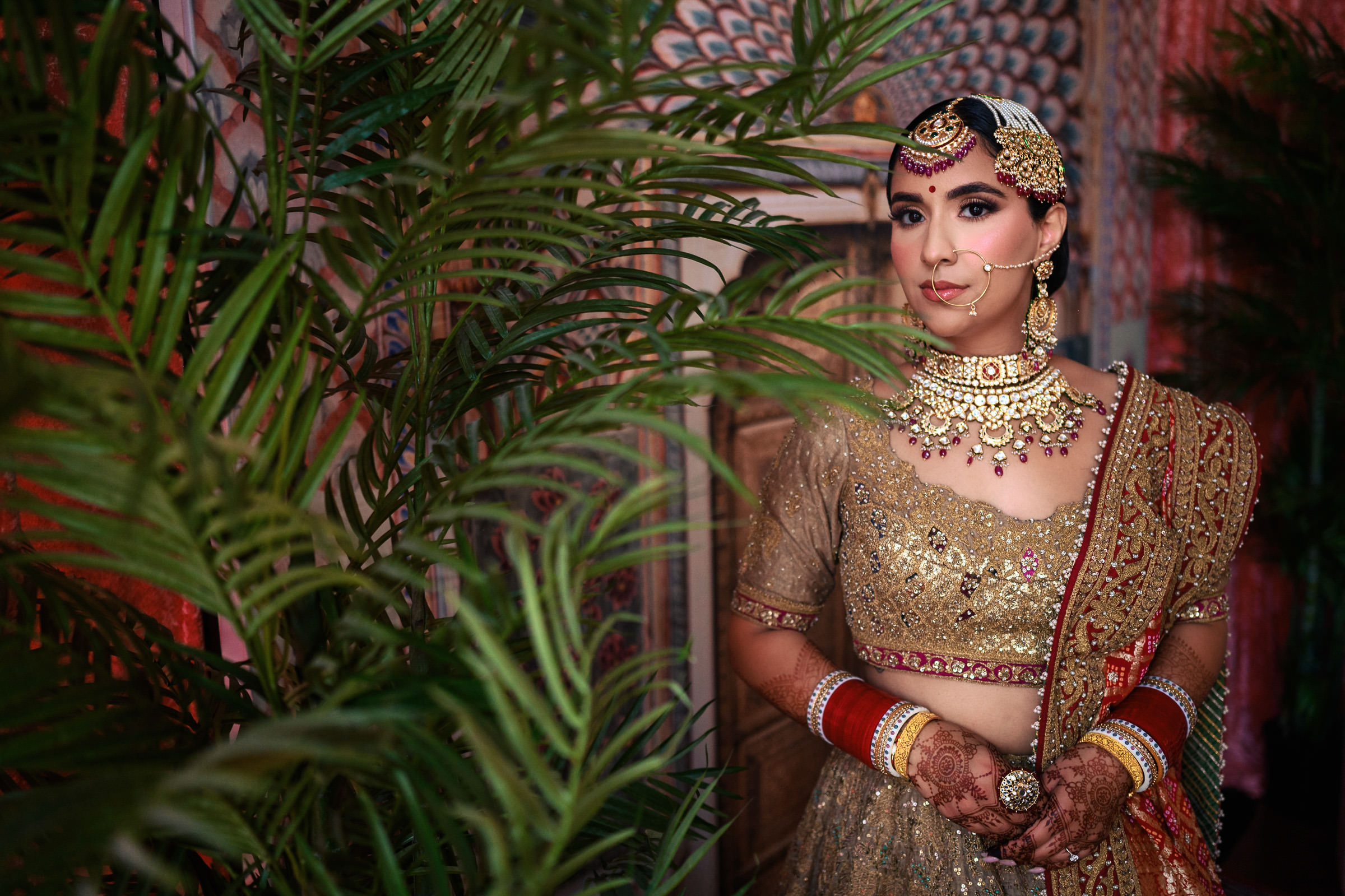 A woman in traditional Indian bridal attire stands near green foliage, capturing one of her favourite wedding photos. She wears a gold lehenga, ornate jewelry, and a nose ring. Her hands are adorned with henna as she poses against a colorful, decorative backdrop.