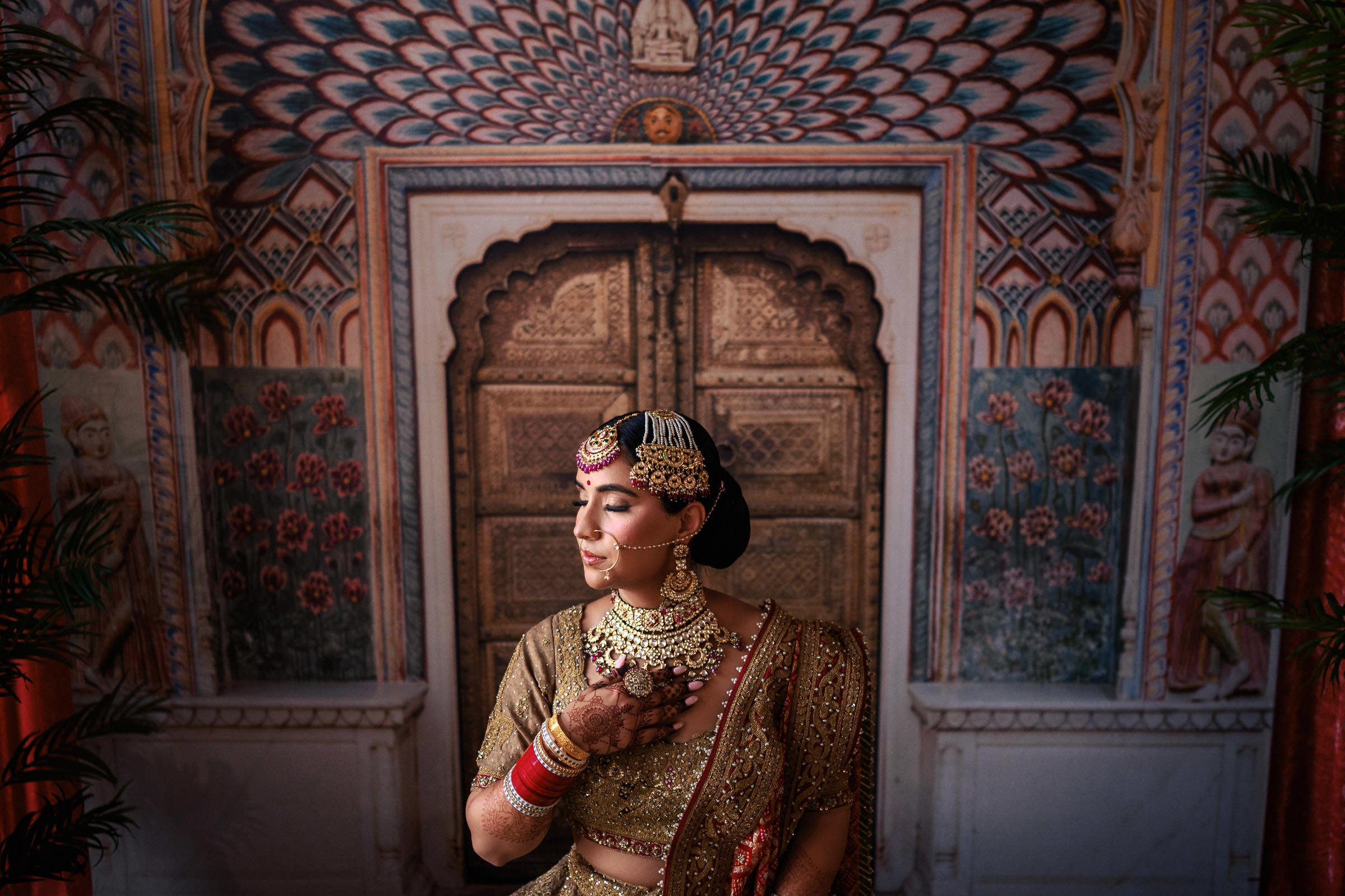 A woman in traditional Indian bridal attire stands with eyes closed and a serene expression, capturing one of her favorite wedding photos. She wears ornate jewelry, including a maang tikka and necklace. The ornate background features a detailed pattern and an arched doorway.