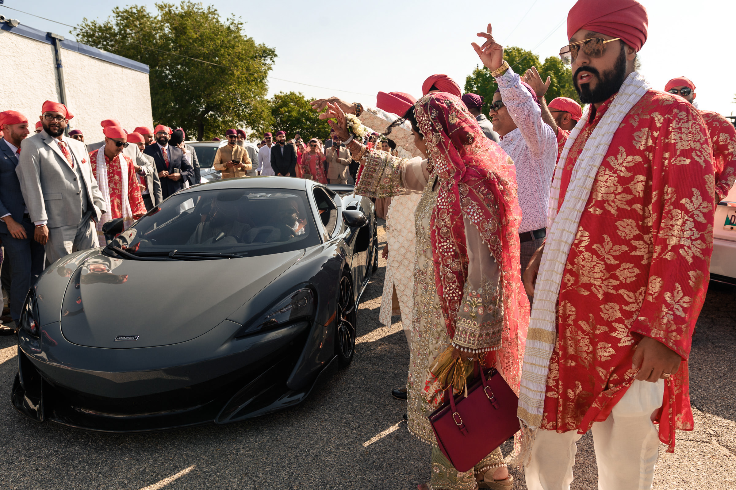 A group of people, dressed in traditional South Asian attire and red turbans, pose for favorite wedding photos near a sleek black sports car. A central figure gestures towards the vehicle as trees and a building provide a picturesque background.