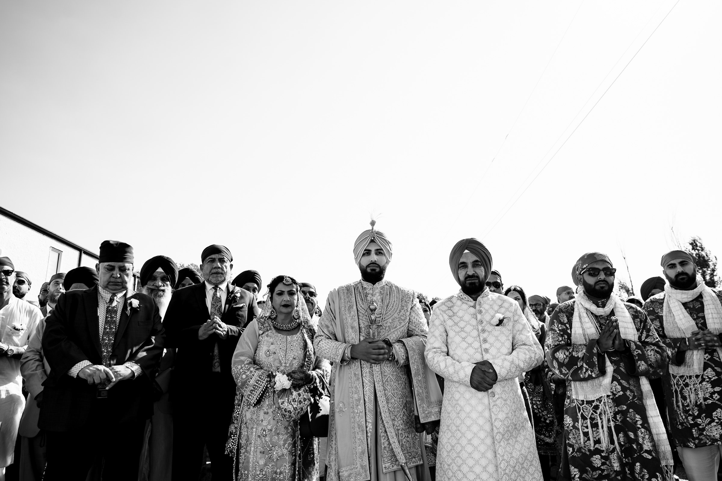 A group of people stands outdoors; some wear traditional attire like turbans and embellished garments. The central figures, captured in one of my favorite wedding photos, appear to be part of a ceremony. The mood is solemn and respectful under a clear sky. Black and white photo.