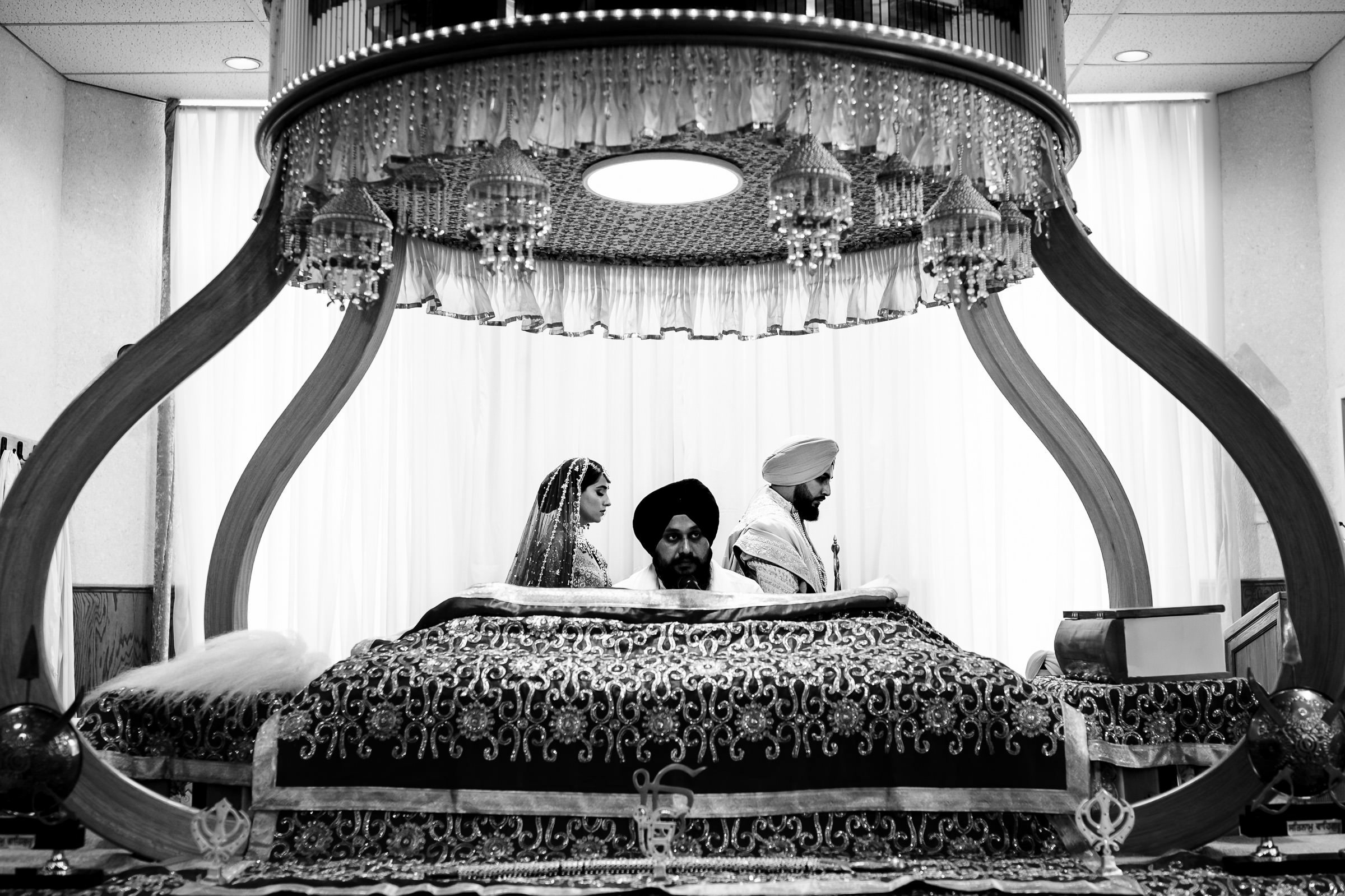 A black and white image captures one of my favorite wedding photos, depicting a Sikh ceremony. The couple sits on the left under an ornate canopy; the groom dons a turban. A person is seated at the richly decorated altar, while elegant drapery gracefully hangs in the background.