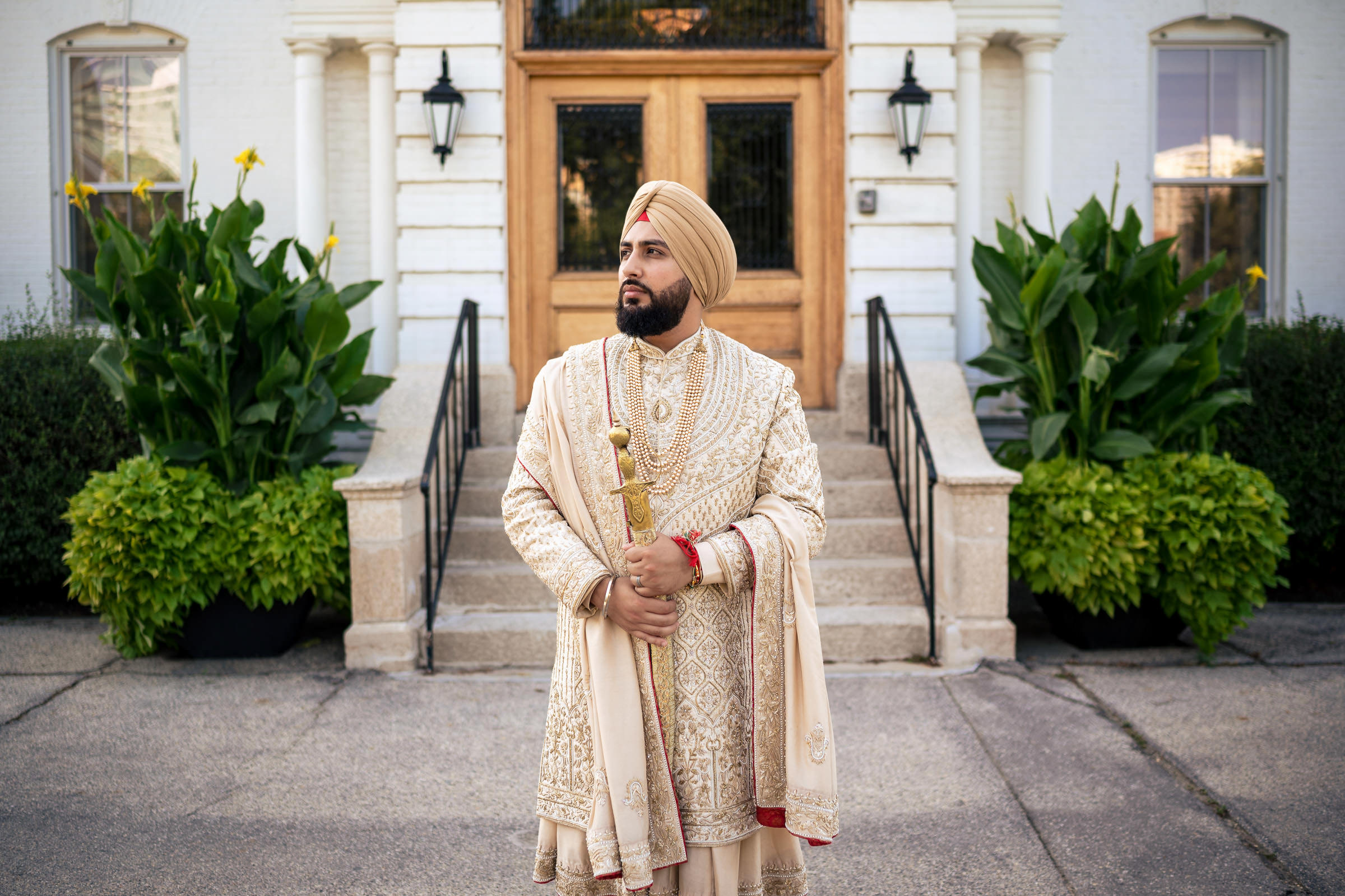 A person wearing an ornate beige sherwani and turban holds a ceremonial sword, captured in one of my favorite wedding photos. They stand before a building adorned with stairs and lush green plants, while the large wooden doors showcase classic architecture.