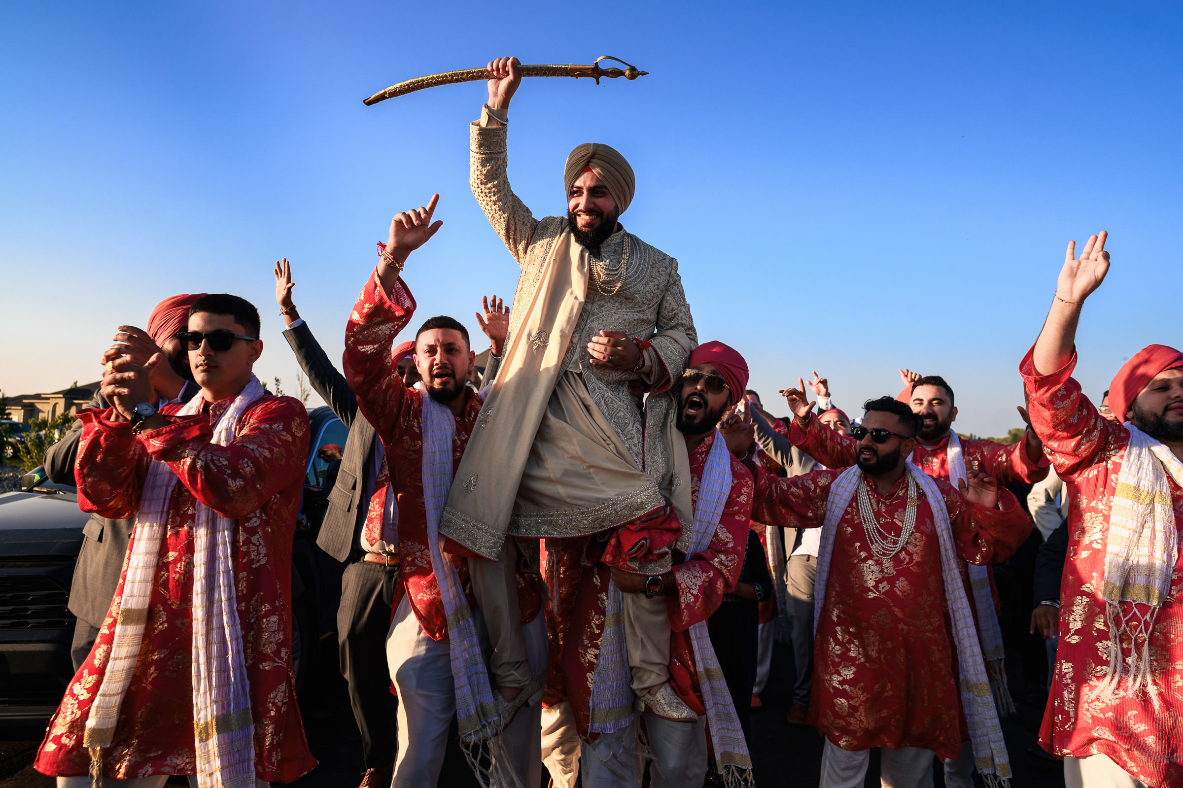 A groom in a traditional outfit is lifted by friends during a lively wedding procession, captured in one of the favorite wedding photos. Holding a decorated stick, he is surrounded by cheerful faces and raised hands against a clear blue sky, while men don red and gold attire.