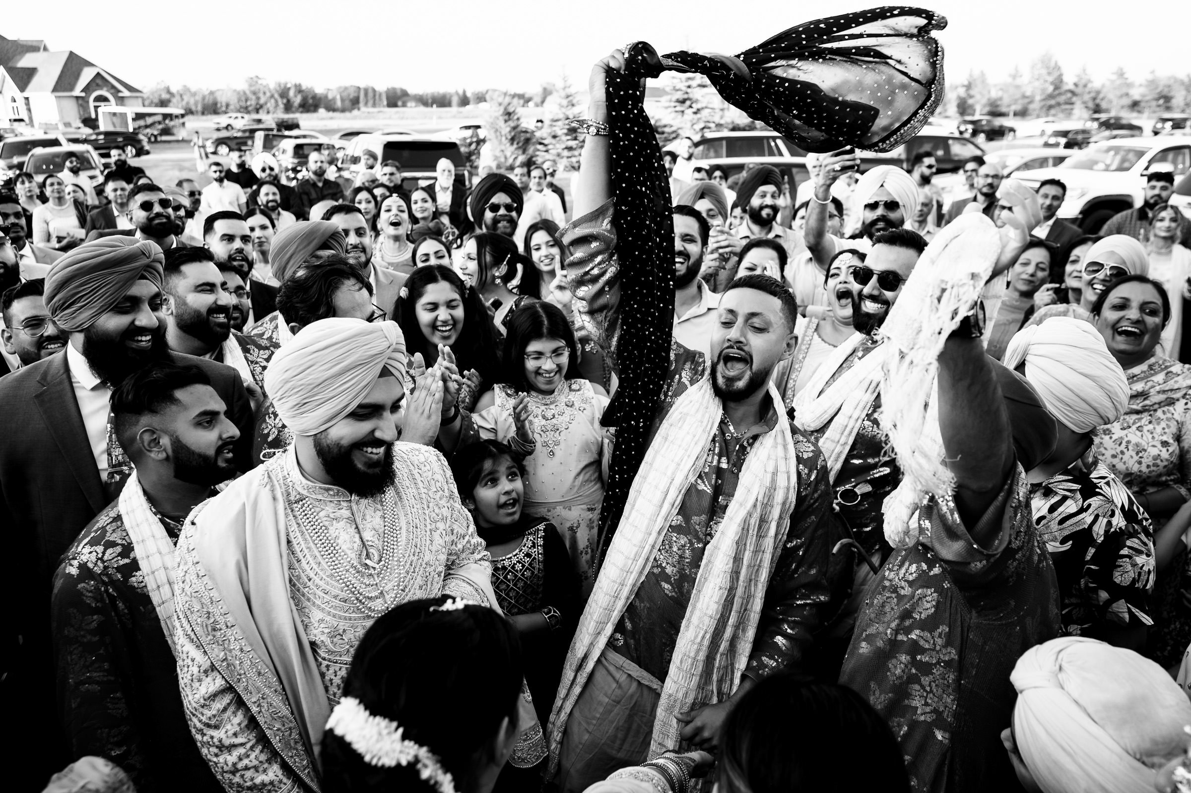 A joyful outdoor celebration unfolds, reminiscent of favorite wedding photos, featuring a crowd in traditional attire. A man energetically raises a cloth while others smile and clap. The scene captures a festive, multicultural gathering with vibrant energy in black and white.