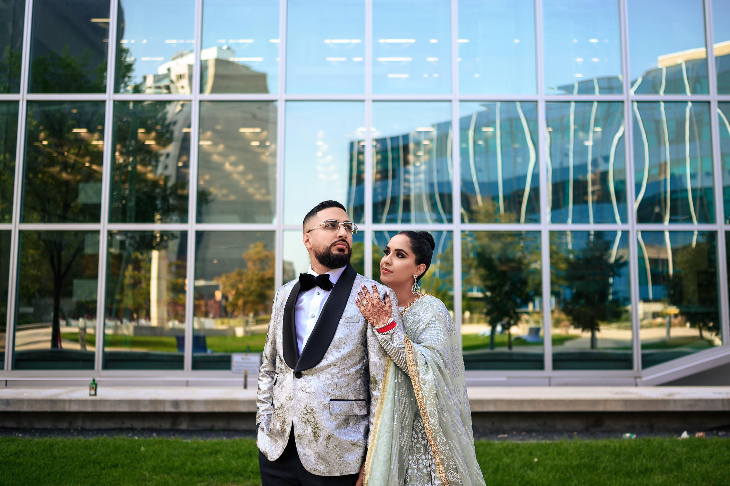 A couple stands in front of a modern glass building, capturing one of their favourite wedding photos. The man is in a silver tuxedo with a black bow tie, while the woman dazzles in a light green embroidered dress, her hands adorned with henna. Both gaze into the distance as trees reflect on the windows.