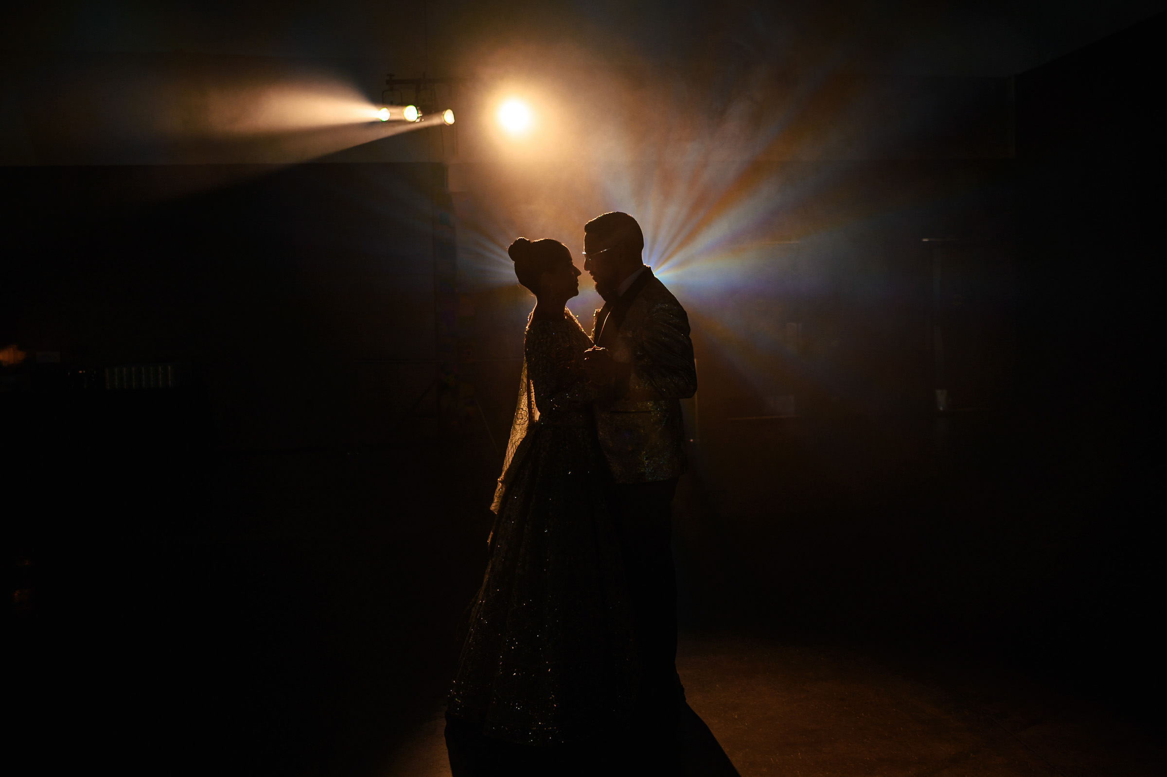 Silhouetted couple dancing closely under dramatic stage lighting, capturing one of their favourite wedding photos. The romantic and intimate atmosphere is enhanced by beams of light radiating in the background.